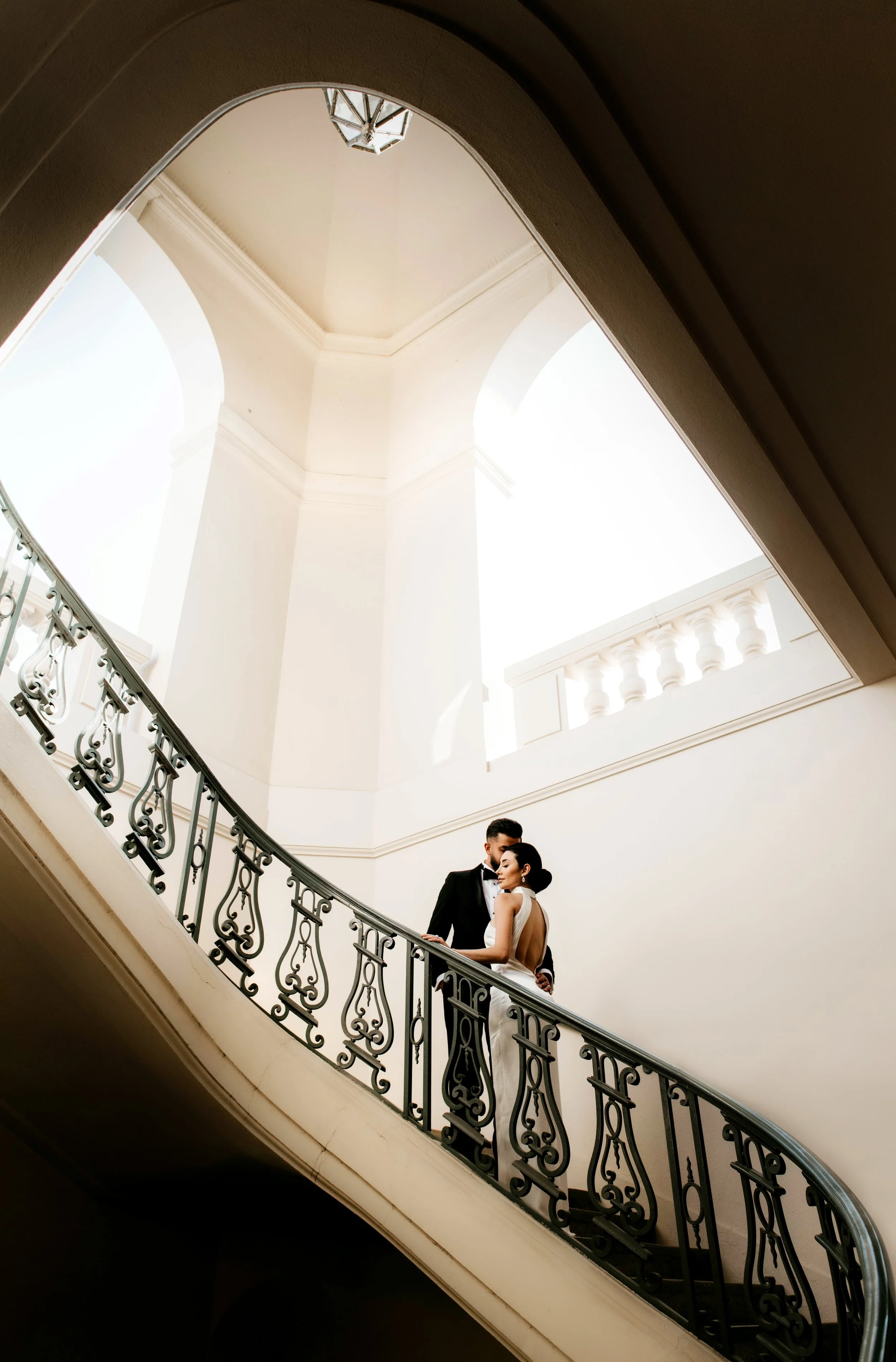 A couple dressed in wedding attire standing on a staircase in a building with high ceilings and white walls, with the groom in a black tuxedo and the bride in a white wedding gown.