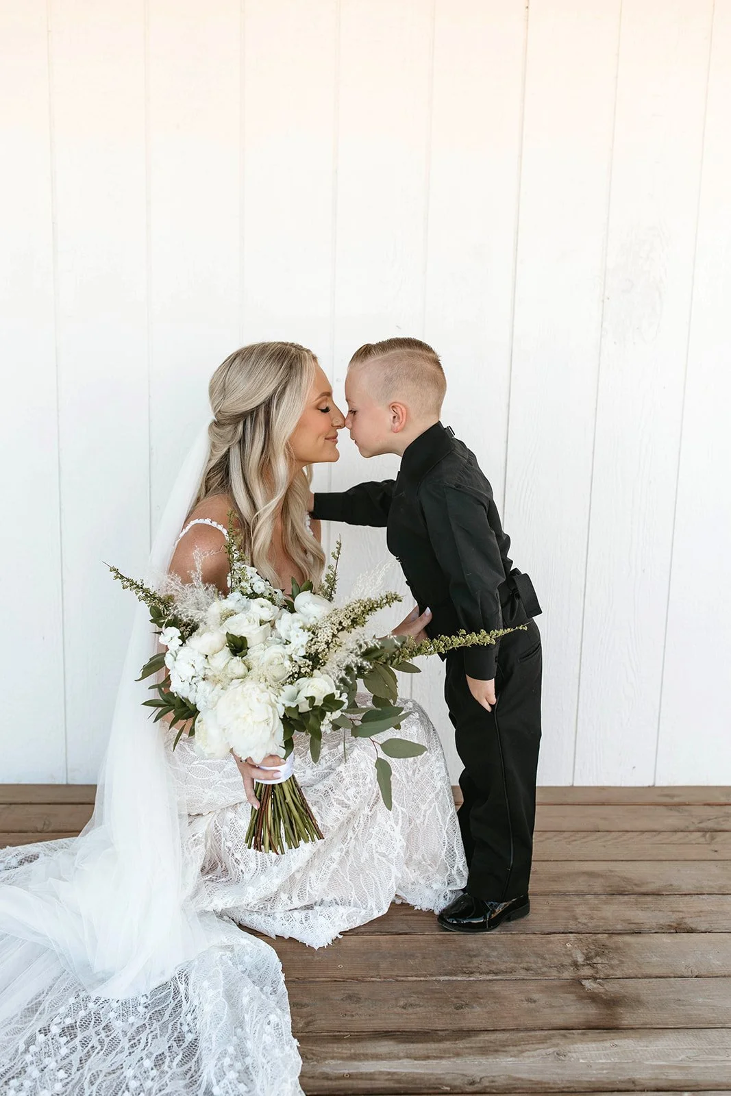 A woman in a wedding dress holding a bouquet of white flowers, and a young boy in a black outfit, touching foreheads against a white wall background.