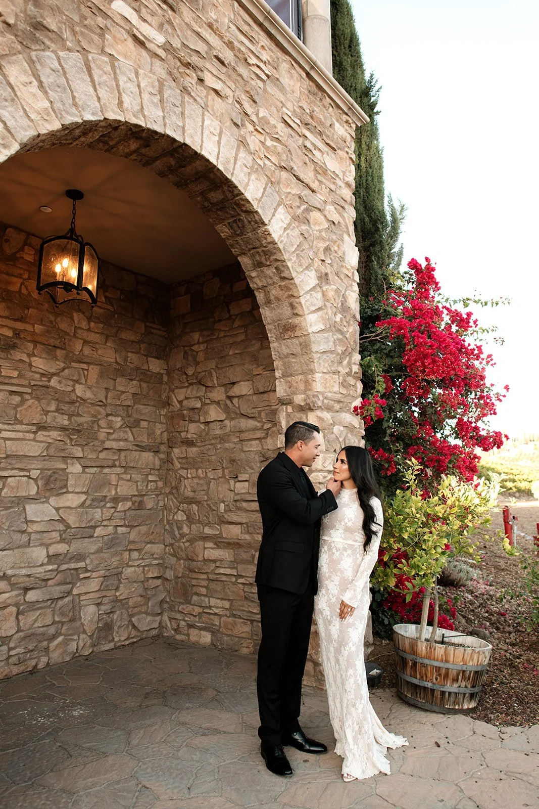 A couple dressed in wedding attire stands close together under an arched stone structure with a hanging lantern, surrounded by blooming pink flowers and a potted plant.