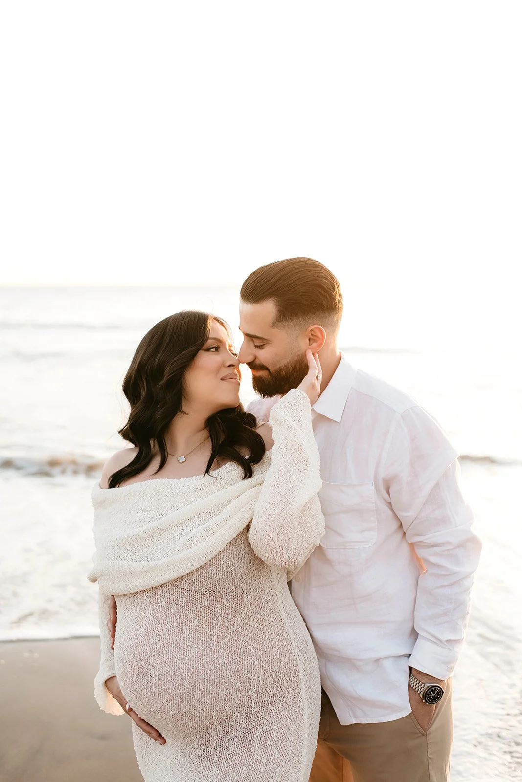 A pregnant woman and a man share a romantic moment on the beach, touching noses, at sunset.