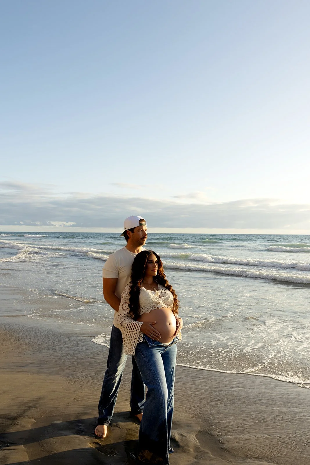 A pregnant woman and a man standing on the beach with the ocean in the background during sunset, the woman is cradling her belly
