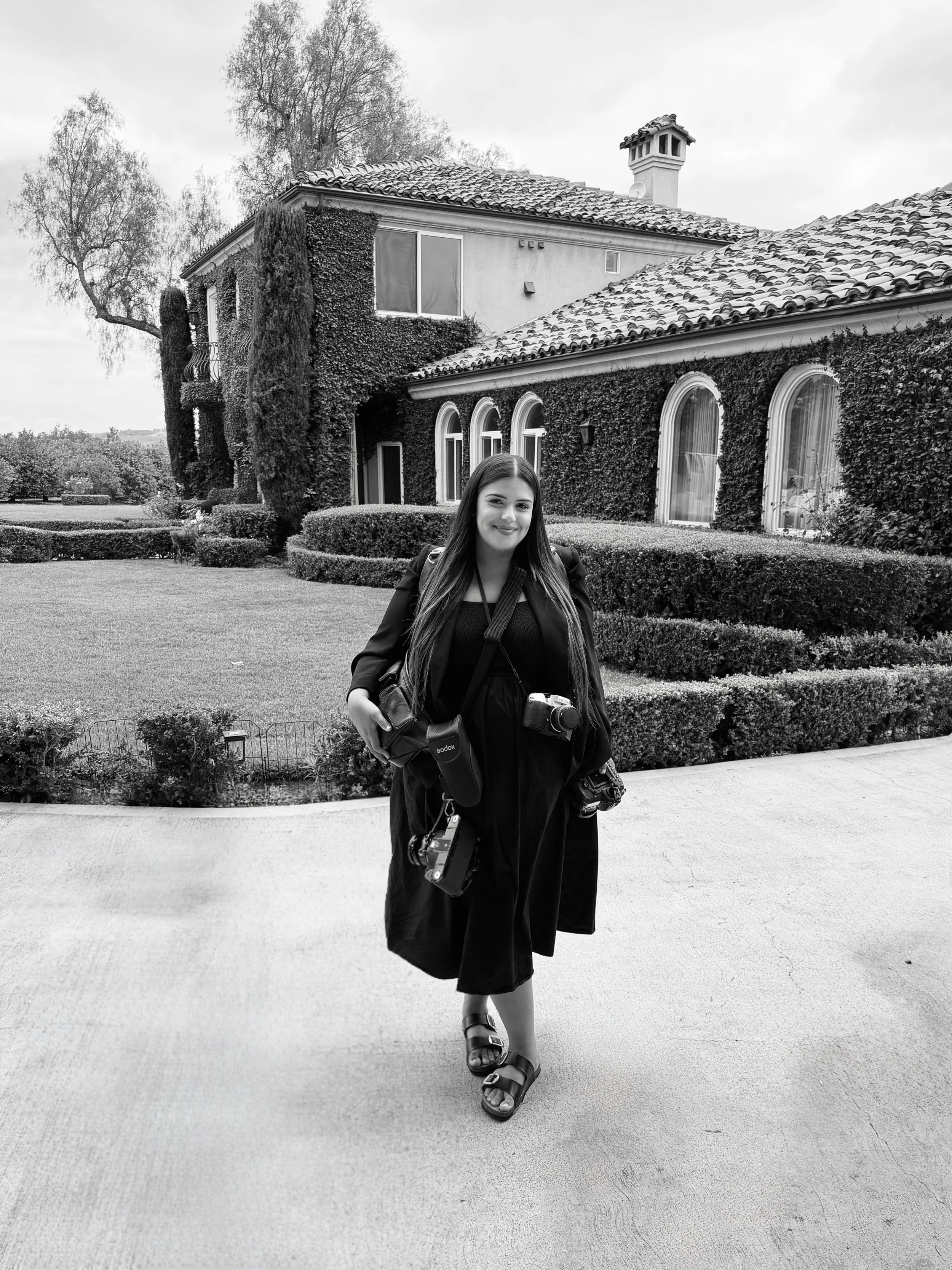A woman with long hair wearing a black dress and sandals, carrying a camera and a camera bag, walking on a pathway in front of a large house covered in ivy, with manicured bushes and trees in the background.