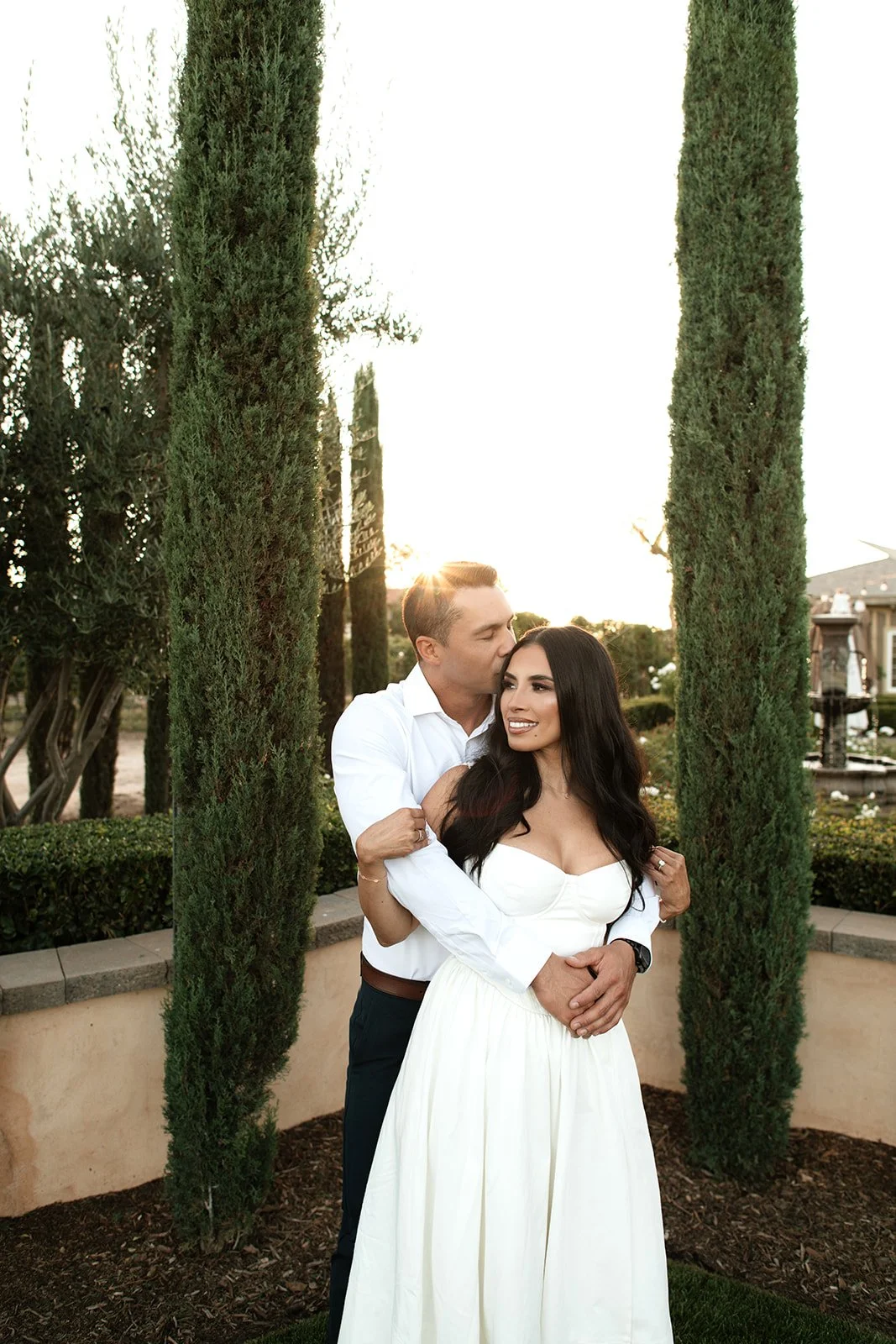 A couple embracing outdoors during sunset, surrounded by tall cypress trees and a garden with a fountain in the background.
