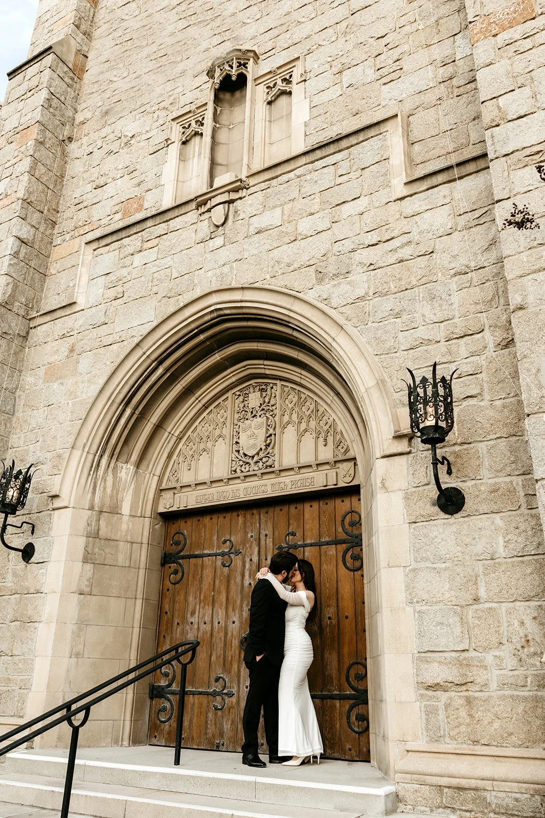 A couple dressed in wedding attire kissing in front of a large wooden door with stone archway at a historic stone building, with black iron lanterns on each side.