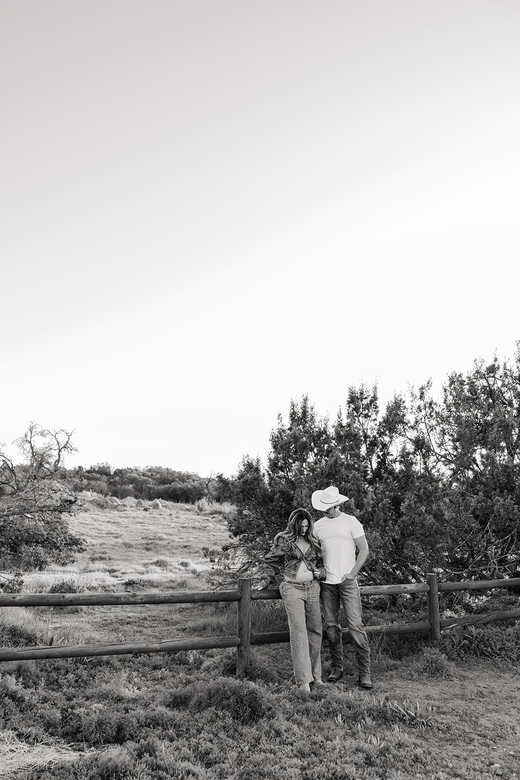 A black and white photo of a couple standing by a wooden fence in a natural landscape with trees and open field, the woman looking at her pregnant belly and the man wearing a hat looking at her.