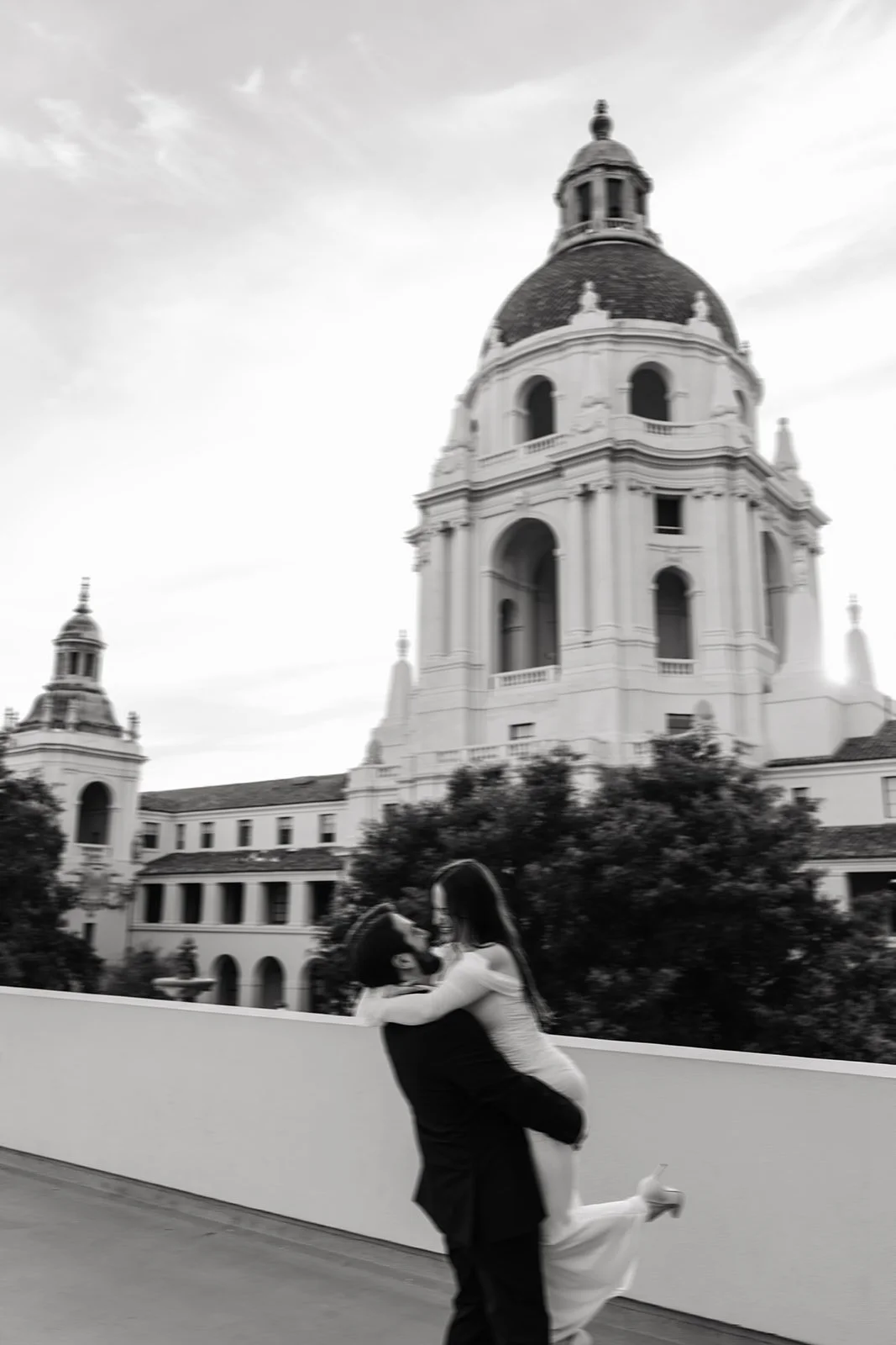 A couple embracing and kissing outdoors with a large historic European-style building with a domed roof in the background.