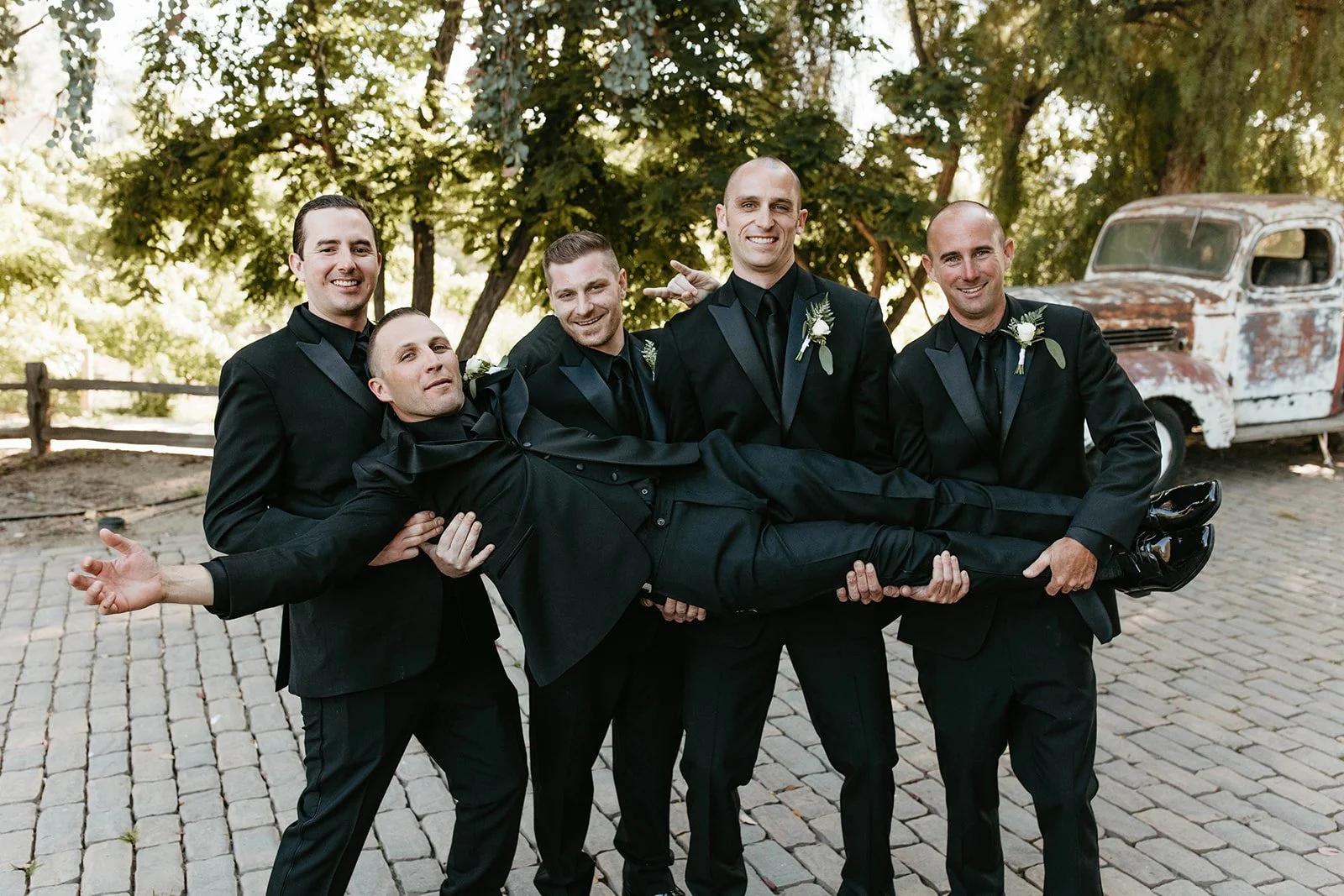Group of six men dressed in black suits and ties, holding a man horizontally in a playful pose outdoors. Backdrop of trees and an old rusty truck.