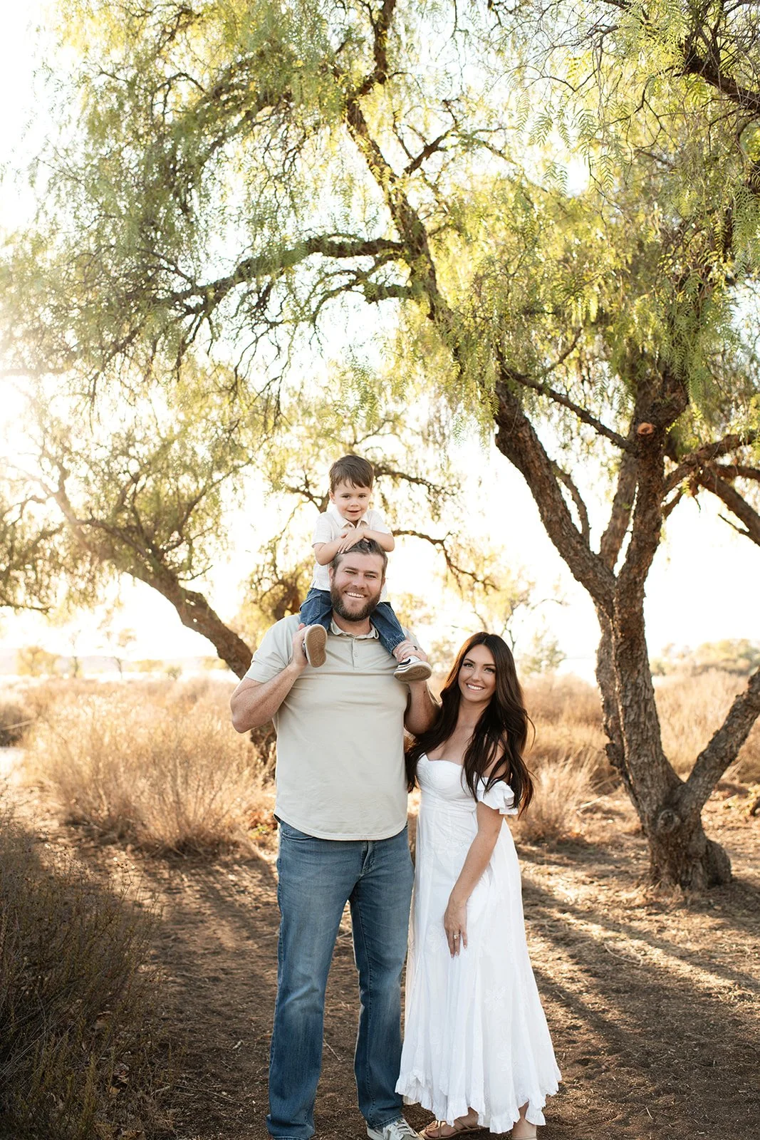 A family of three outdoors on a sunny day, standing in front of trees and dry grass. The father is carrying a young boy on his shoulders, and the mother stands beside them, smiling.