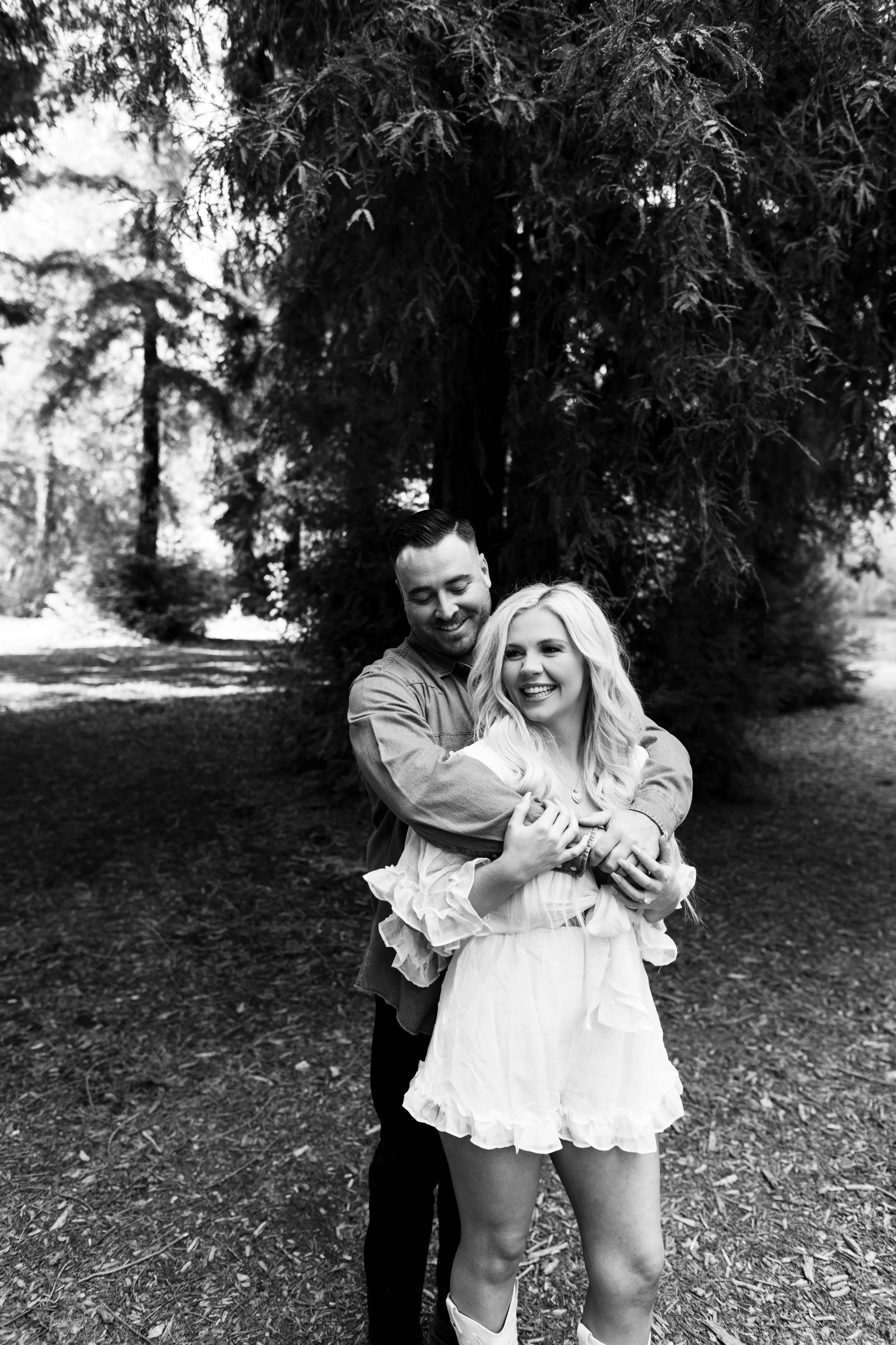 A black-and-white photo of a smiling woman with long, wavy hair standing in front of a tall tree, with a man embracing her from behind, in a park setting with grass and other trees visible in the background.