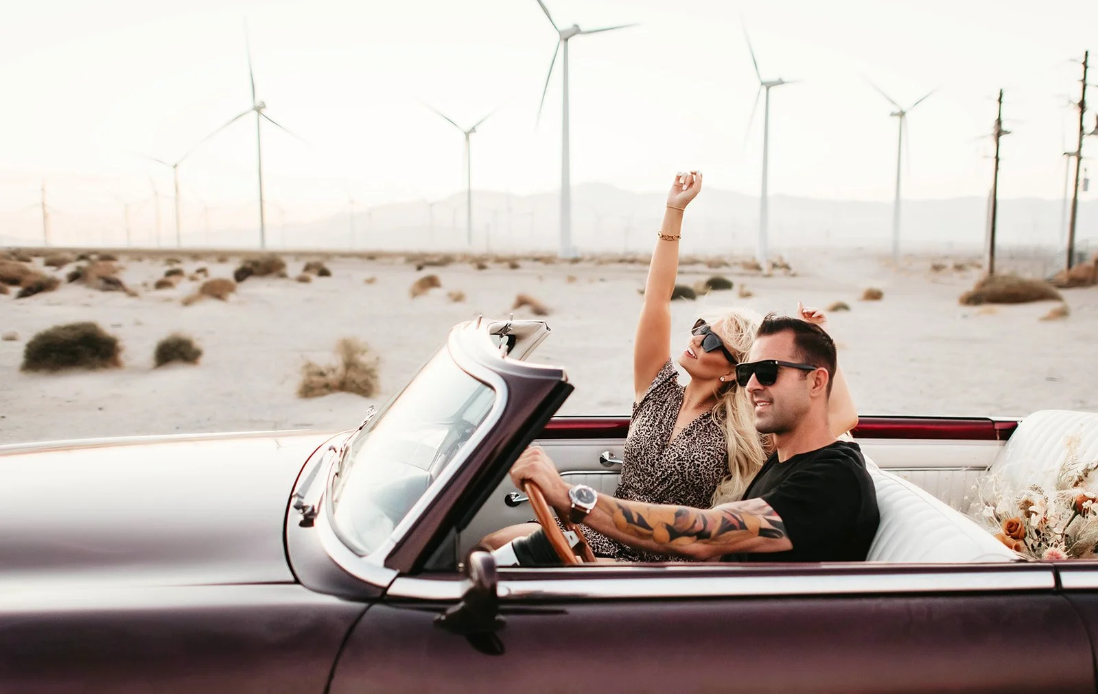 A couple enjoying a ride in a vintage convertible car with a barren desert landscape and wind turbines in the background. The woman is smiling, raising her arms, and wearing sunglasses, while the man is driving and also wearing sunglasses.