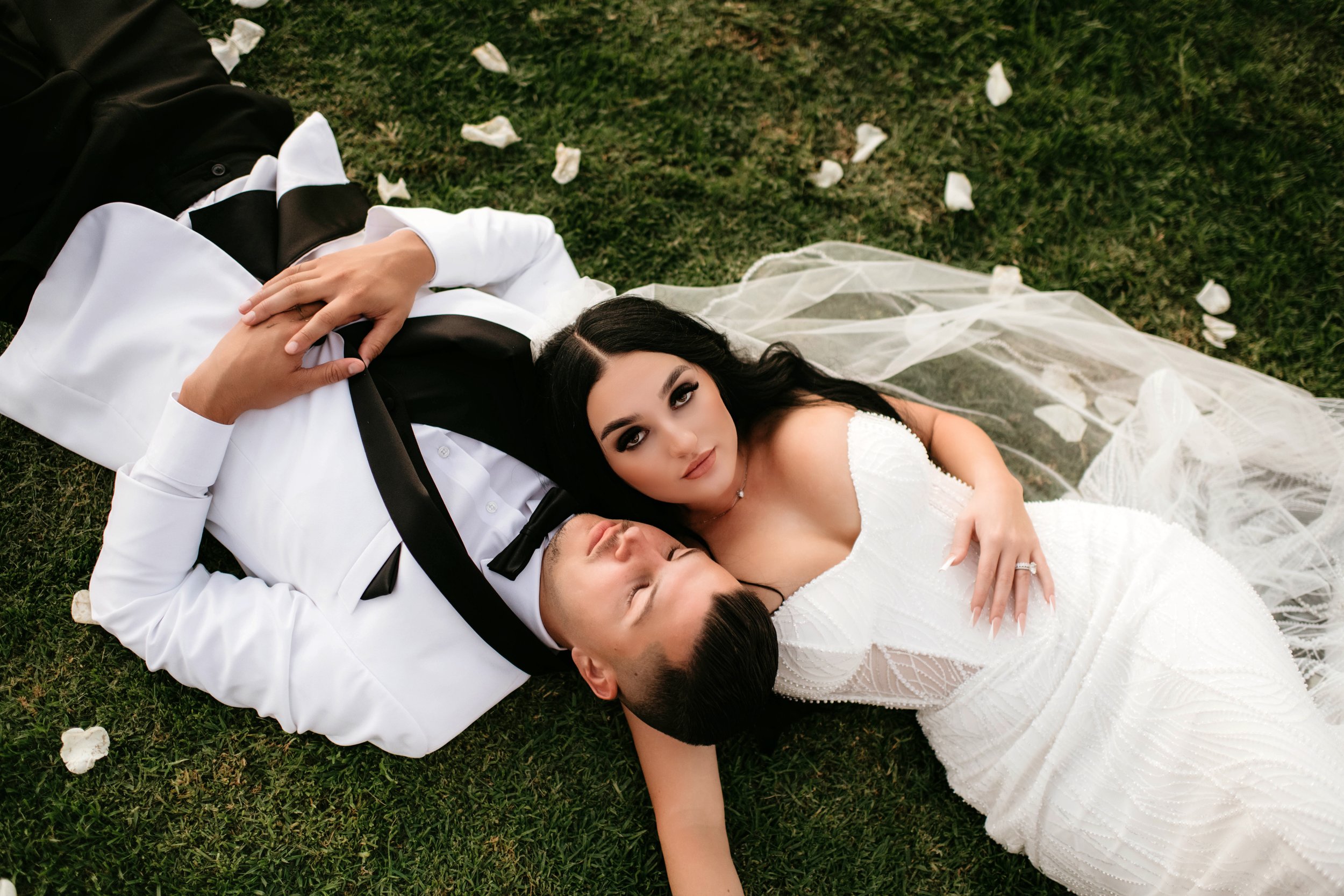 A bride and groom laying on grass, surrounded by flower petals, with the groom wearing a tuxedo and the bride in a white wedding dress