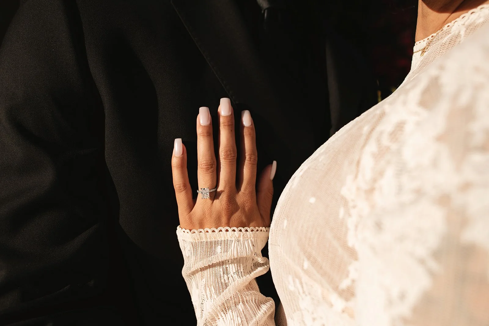 Close-up of a woman's left hand resting on a man's black suit. The woman is wearing a diamond engagement ring and has manicured nails. She is dressed in a cream-colored lace dress with a scalloped sleeve edge.