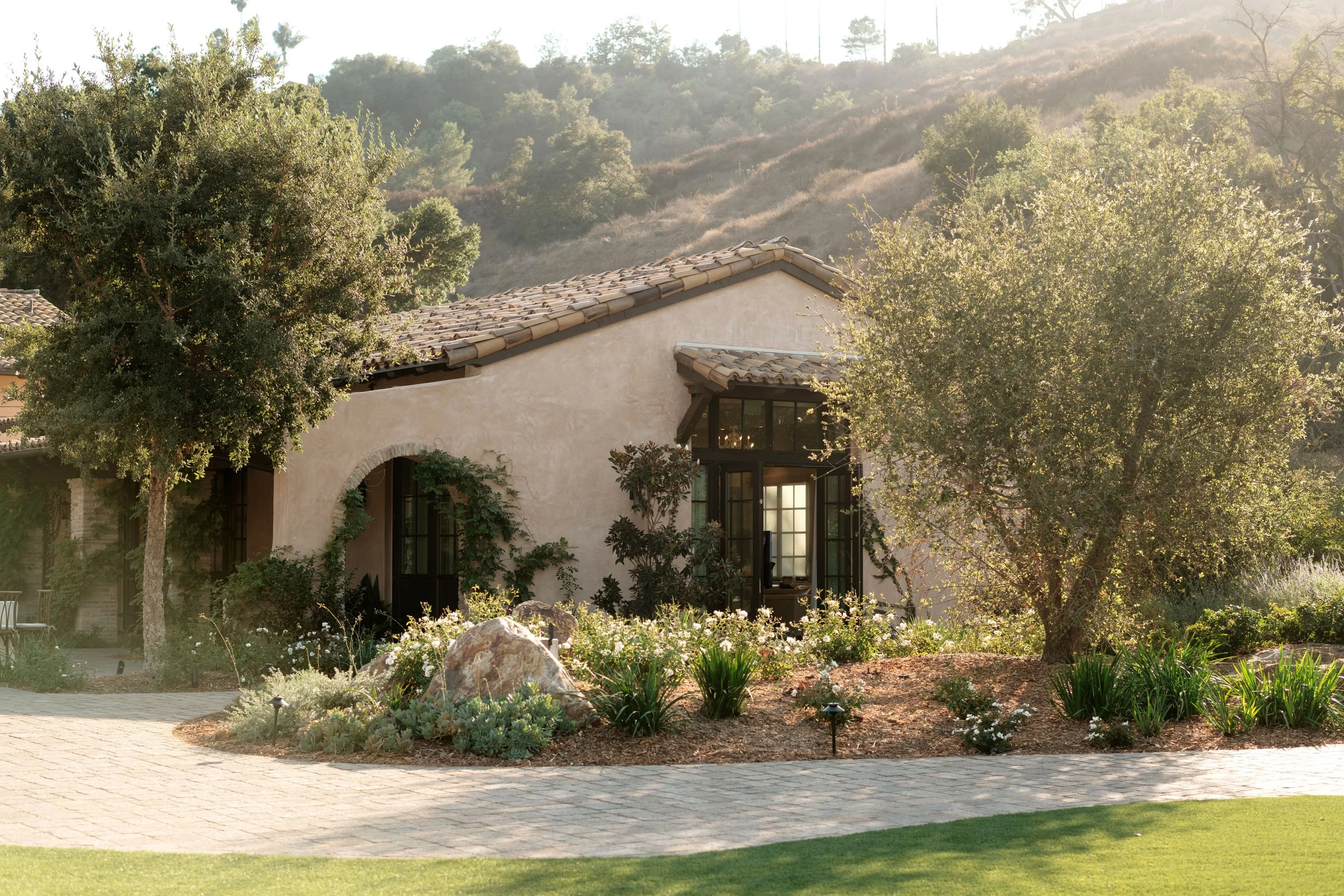 A house with beige stucco walls and a terra cotta tiled roof nestled among trees and shrubs on a sunny day, with a hillside covered in trees in the background.