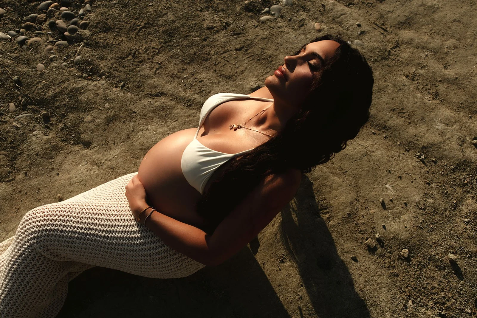 A pregnant woman lying on sandy ground, wearing a white bikini top and crochet pants, with her eyes closed and a peaceful expression.