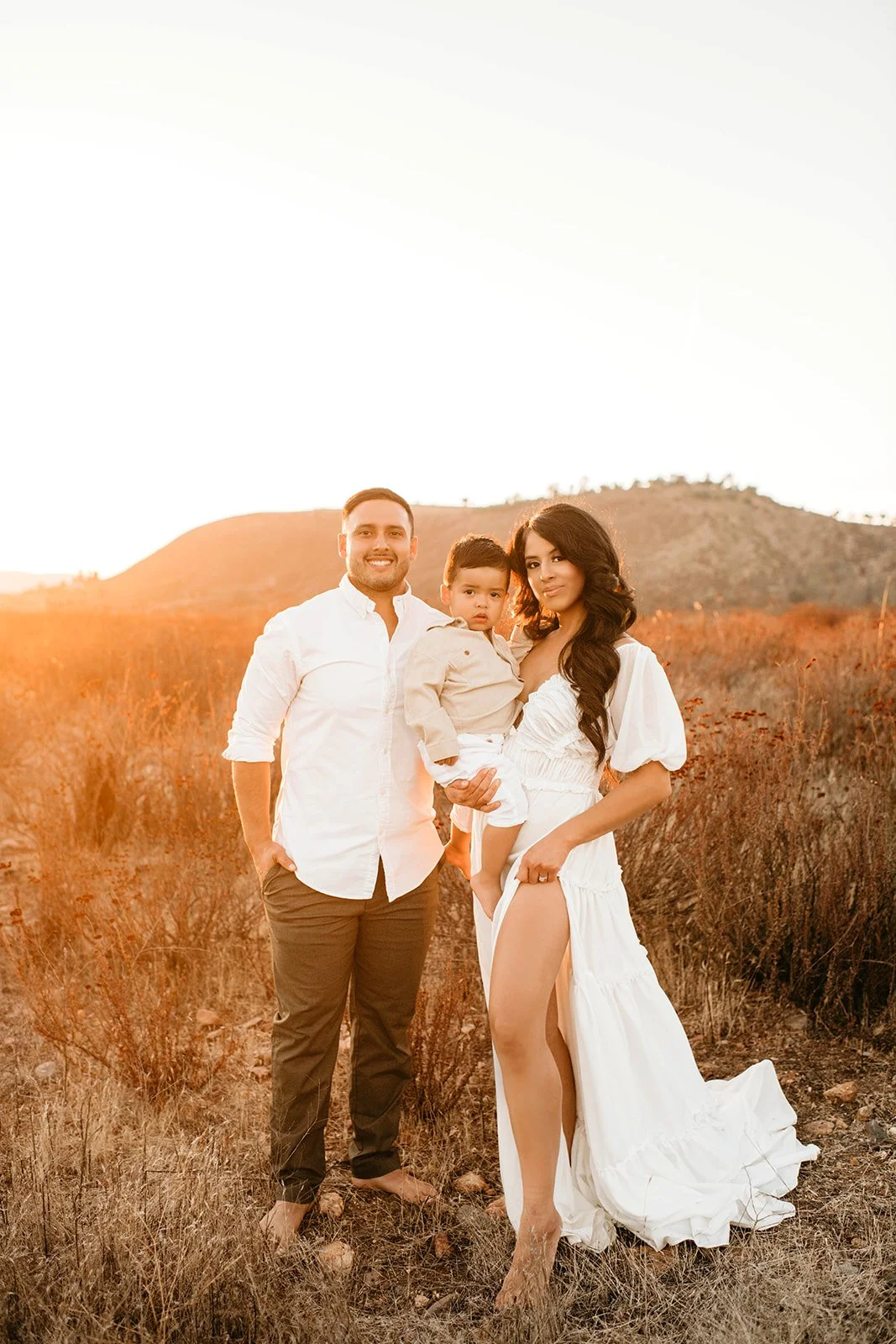 A family of three standing outdoors during sunset, with dry grass and hills in the background. The man is smiling, wearing a white shirt and brown pants, the woman holds a young boy, and she is dressed in a white dress.