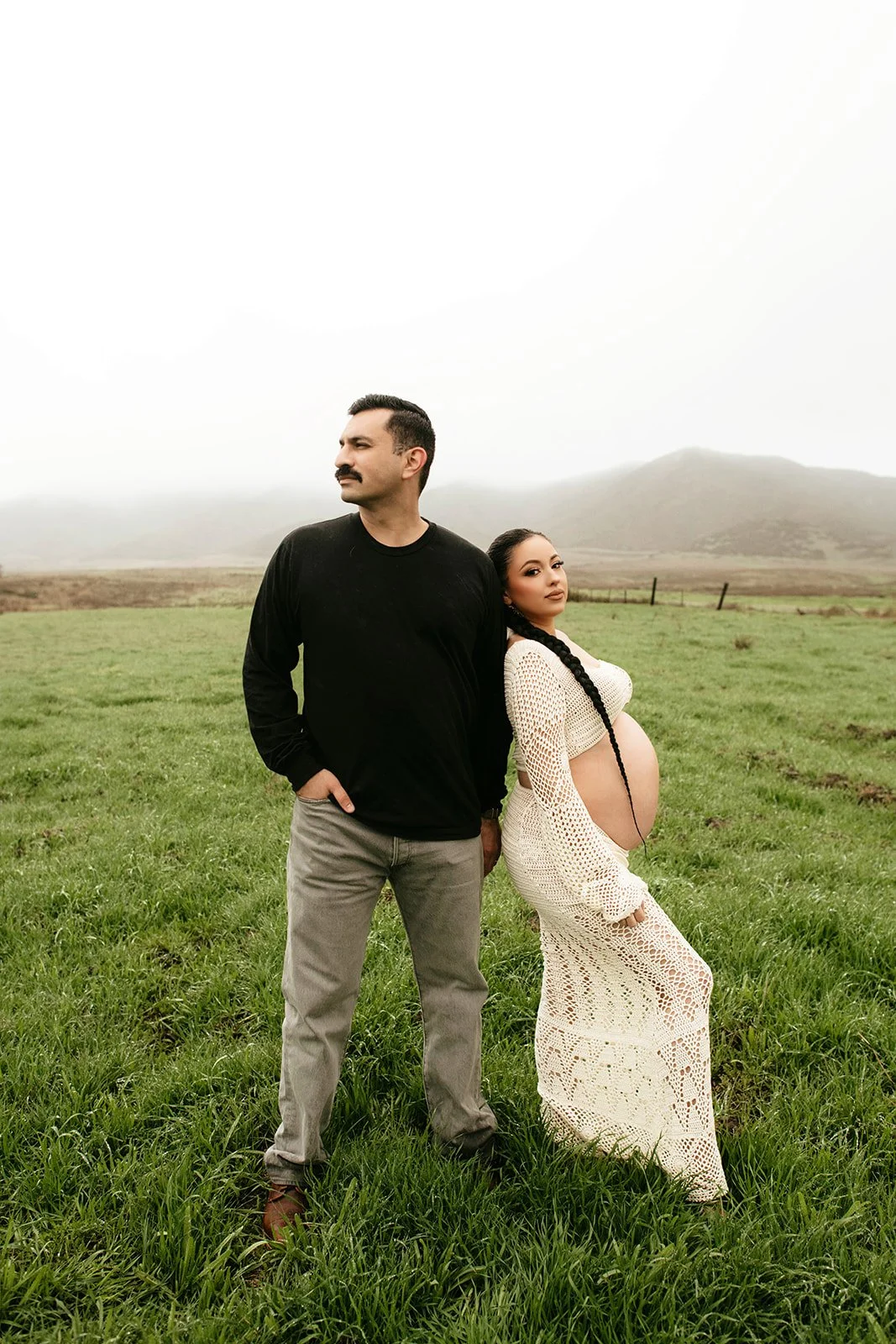 A man and a pregnant woman standing on a grassy field with hills in the background, the woman leaning her head on the man's shoulder.
