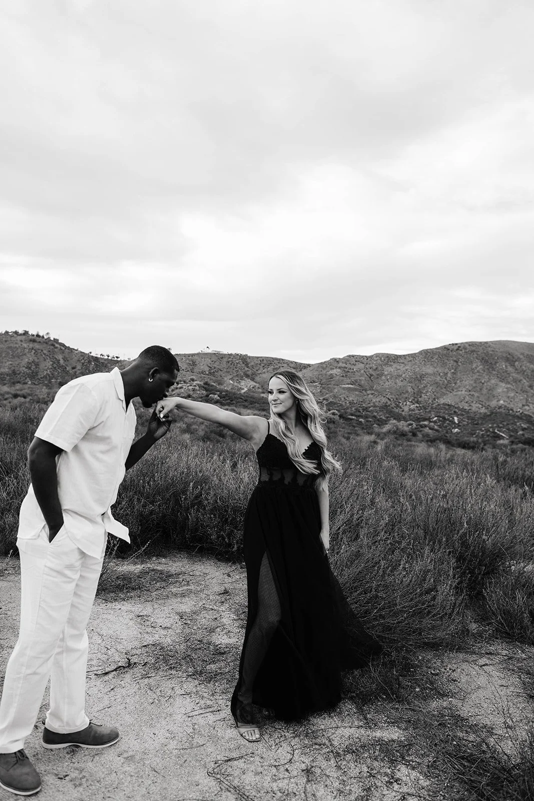 A black and white photo of a woman in a long black dress with sheer panels, standing outdoors on a dirt path with mountains and cloudy sky in the background, as a man in white outfit kisses her hand.