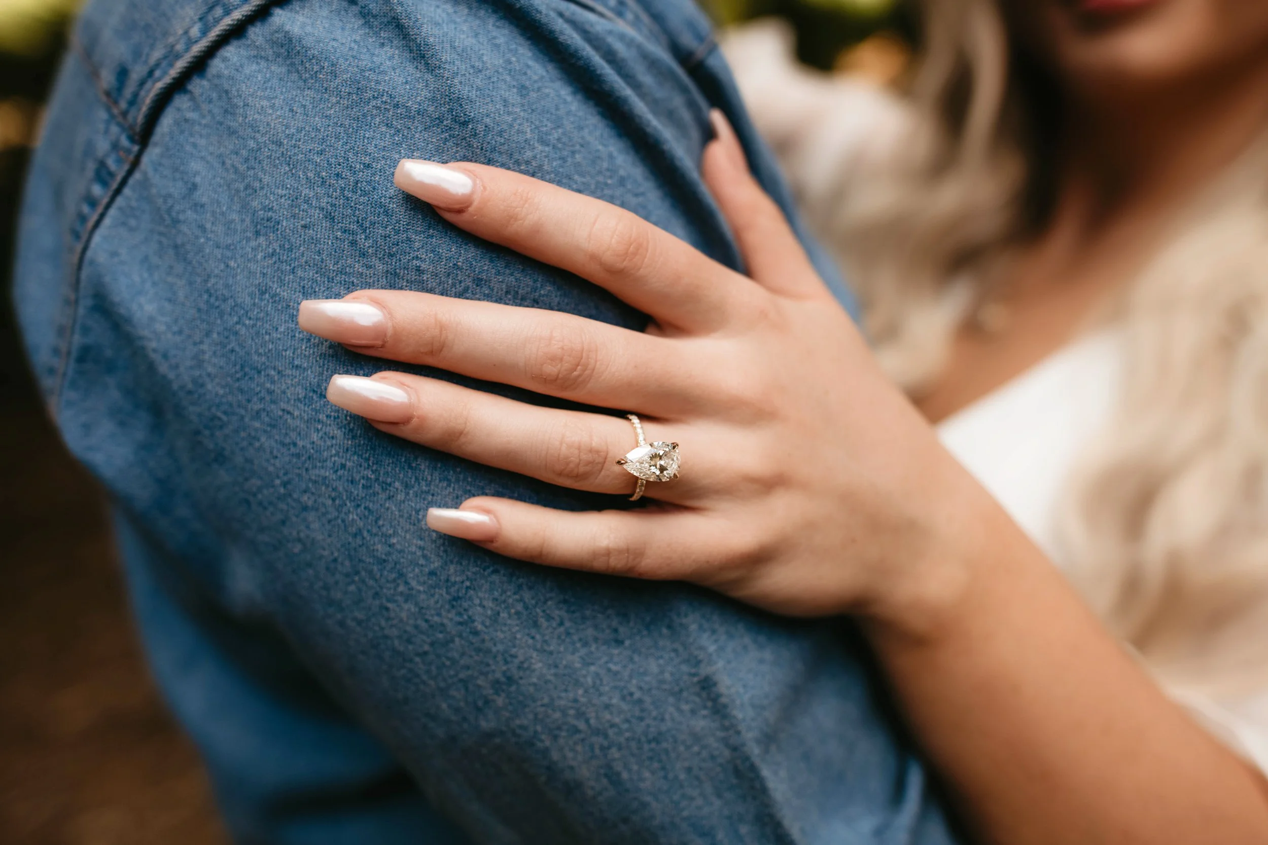 A woman with a large engagement ring on her finger rests her hand on a man's shoulder, who is wearing a blue denim shirt.