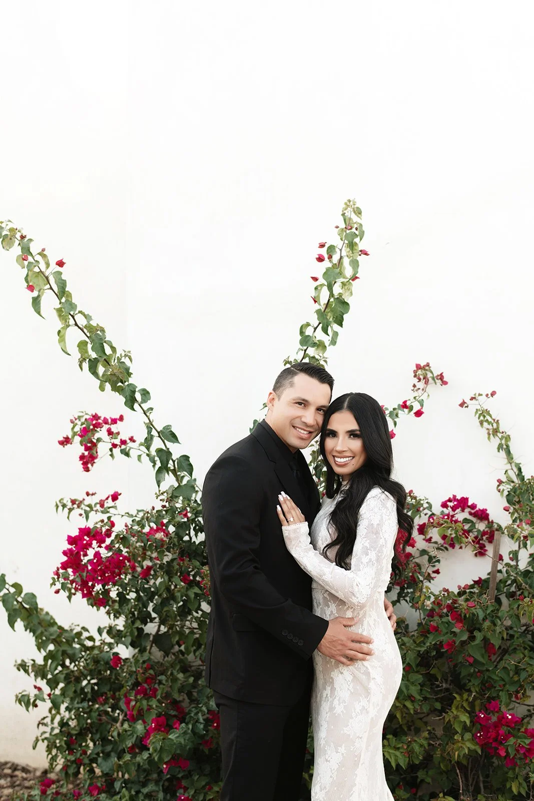 Couple in wedding attire standing close together in front of flowering bushes, smiling.