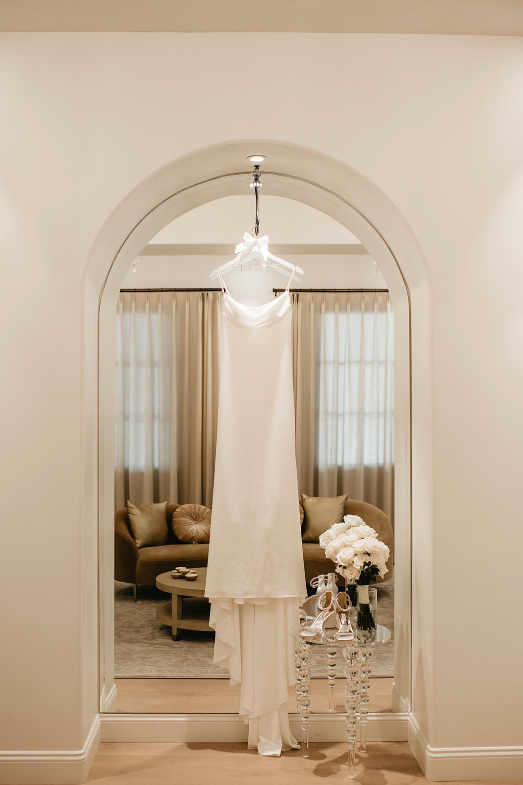 A white wedding dress hanging on a hanger in front of a mirror, with a veil and white high-heeled shoes on a glass table next to a bouquet of white flowers in a black vase, in a room with beige curtains and a beige sofa.