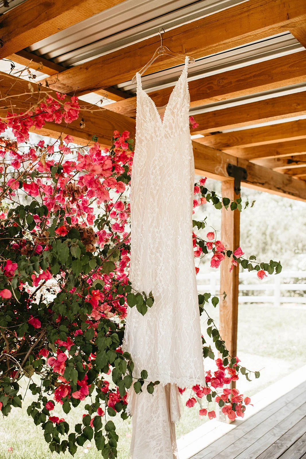 A wedding dress hanging on a hanger from a wooden beam, with pink bougainvillea flowers in the background.