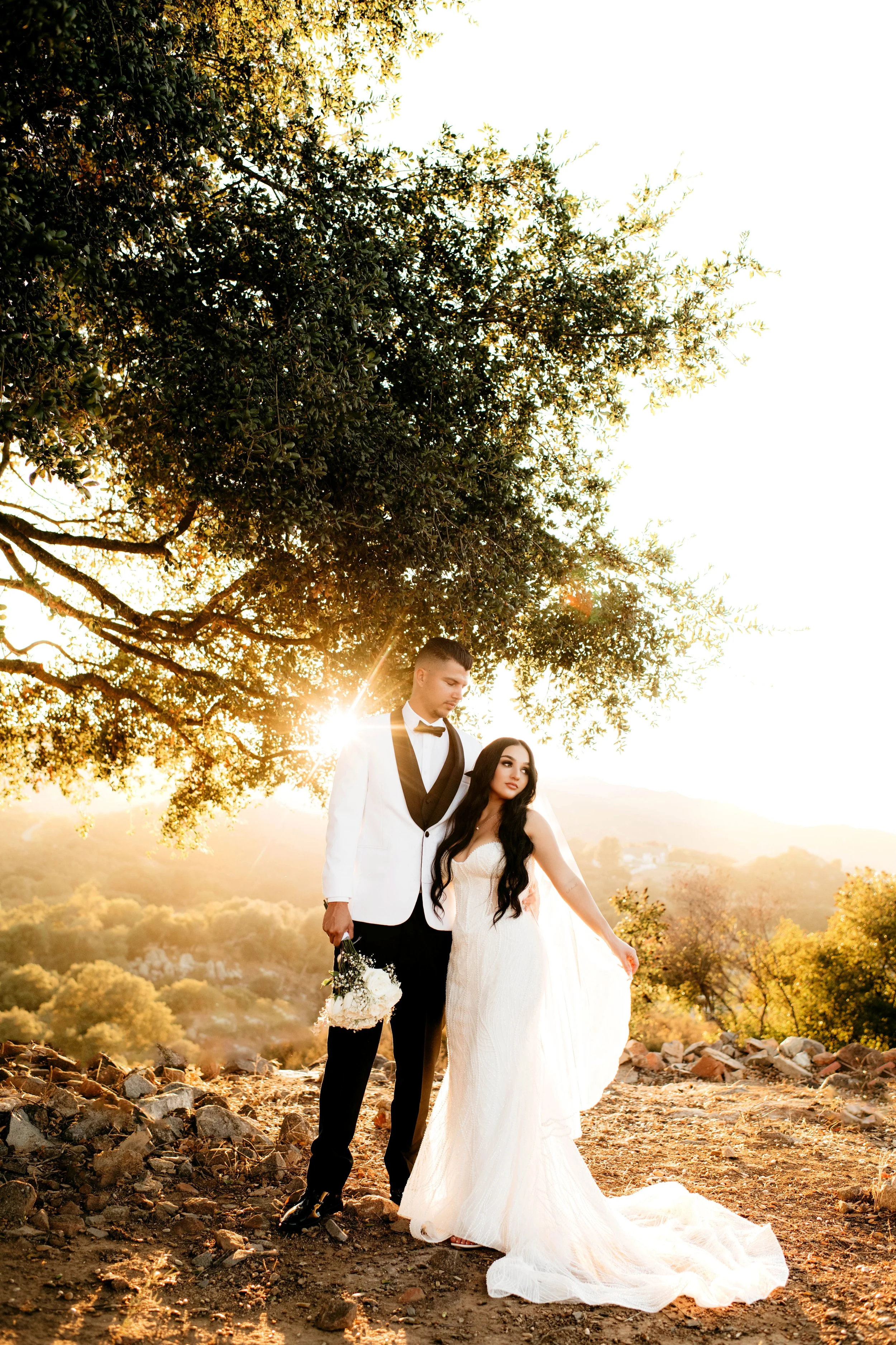 A bride and groom standing outside during sunset, with a large tree overhead and a scenic landscape in the background. The groom wears a white tuxedo with black lapels, and the bride wears a white wedding gown with long dark hair. The sun is shining 