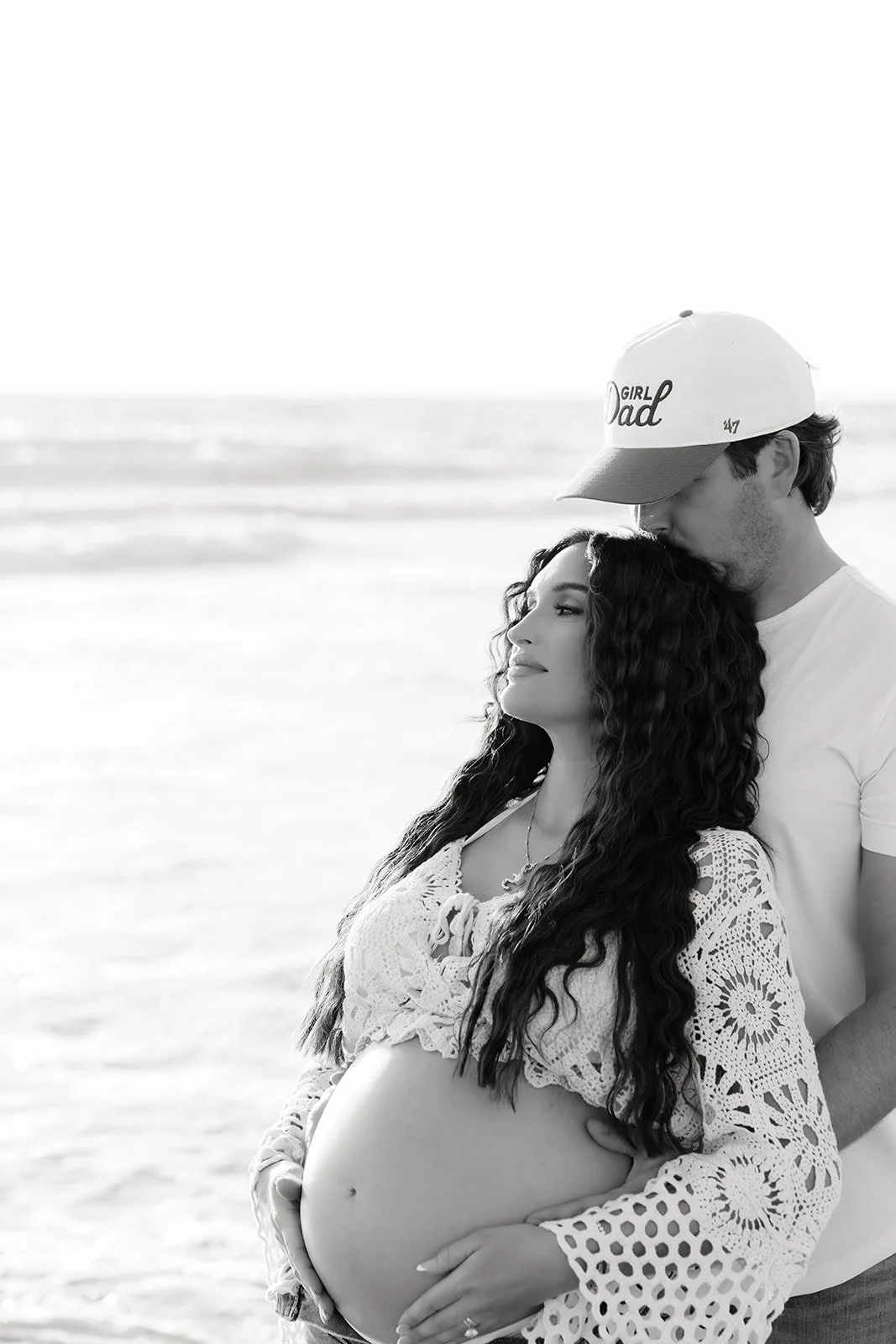 A pregnant woman with long curly hair and a man wearing a cap and white shirt standing on a beach, with the man kissing the woman's forehead.
