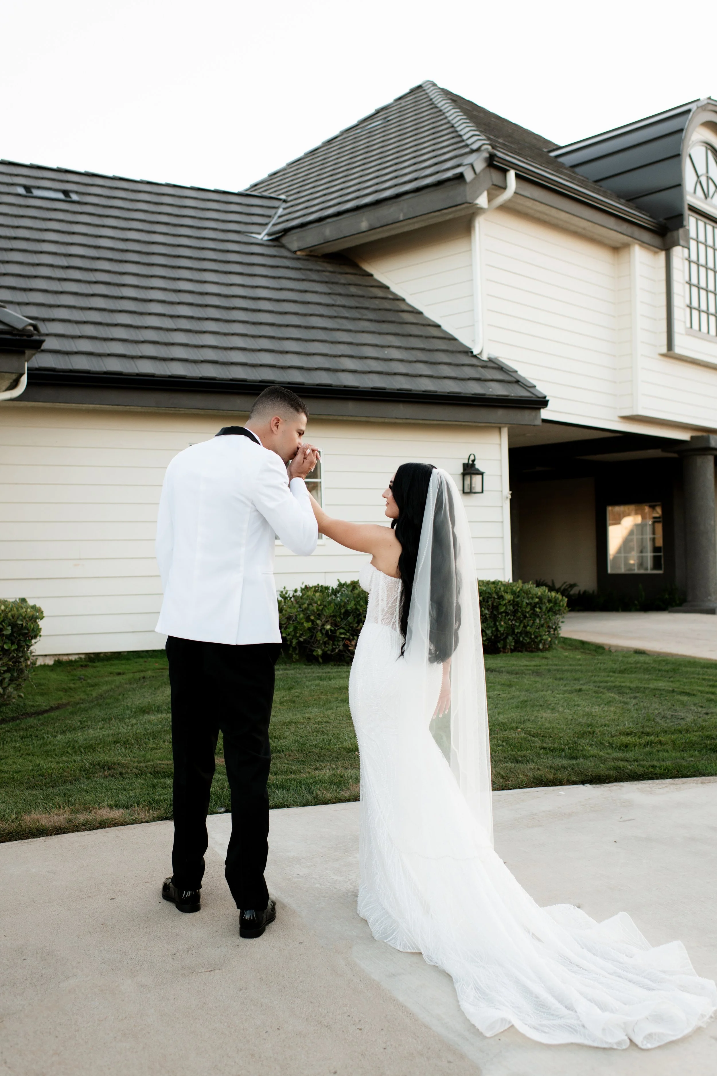 A bride and a groom sharing a moment outside a house, with the groom kissing the bride's hand, both dressed in wedding attire. The bride is in a white wedding gown with a veil, and the groom wears a white jacket with black pants.
