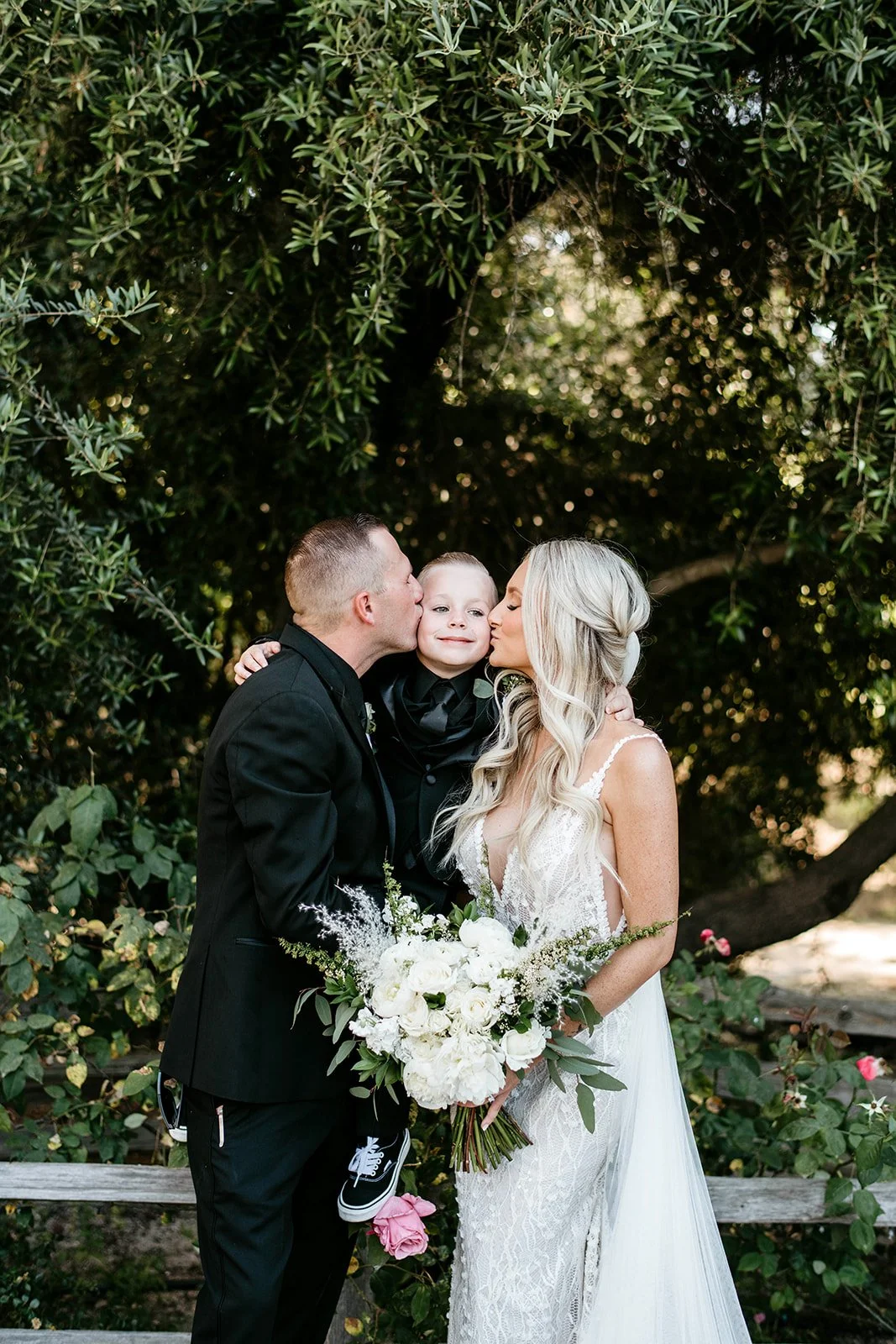 A bride and groom share kisses with a young boy between them, outdoors surrounded by green foliage and trees. The bride holds a large white floral bouquet, and the boy is wearing a black suit with sneakers.