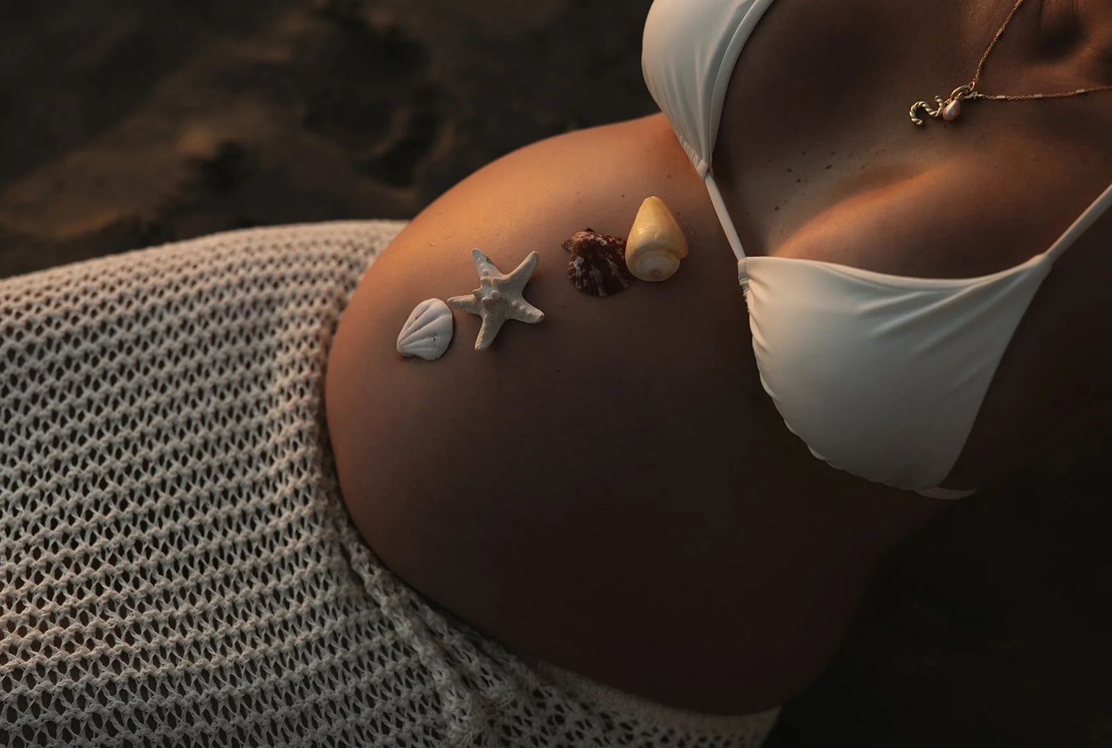 A person's shoulder with seashells and a starfish arranged on it, wearing a white bikini top and a gold necklace, with a knitted garment partially visible.