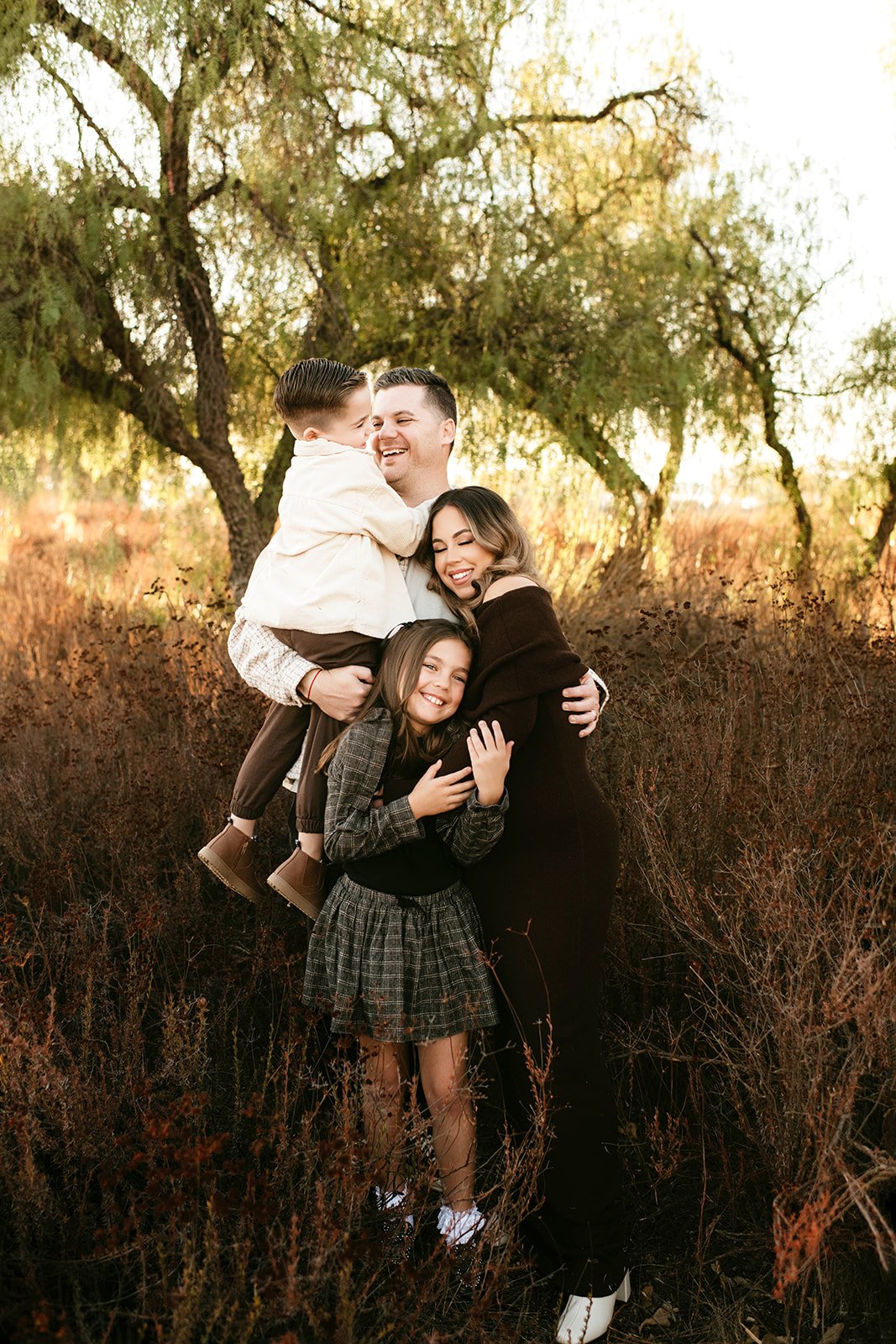 Family of four hugging outdoors in autumn, with trees and dry grass in the background, smiling and enjoying each other's company.