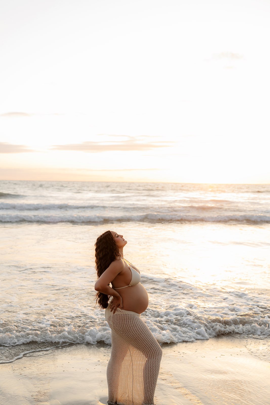 Pregnant woman with long curly hair standing on the beach at sunset, wearing a white bikini top and beige crochet skirt, with her eyes closed, facing the ocean, and the waves gently touching her feet.