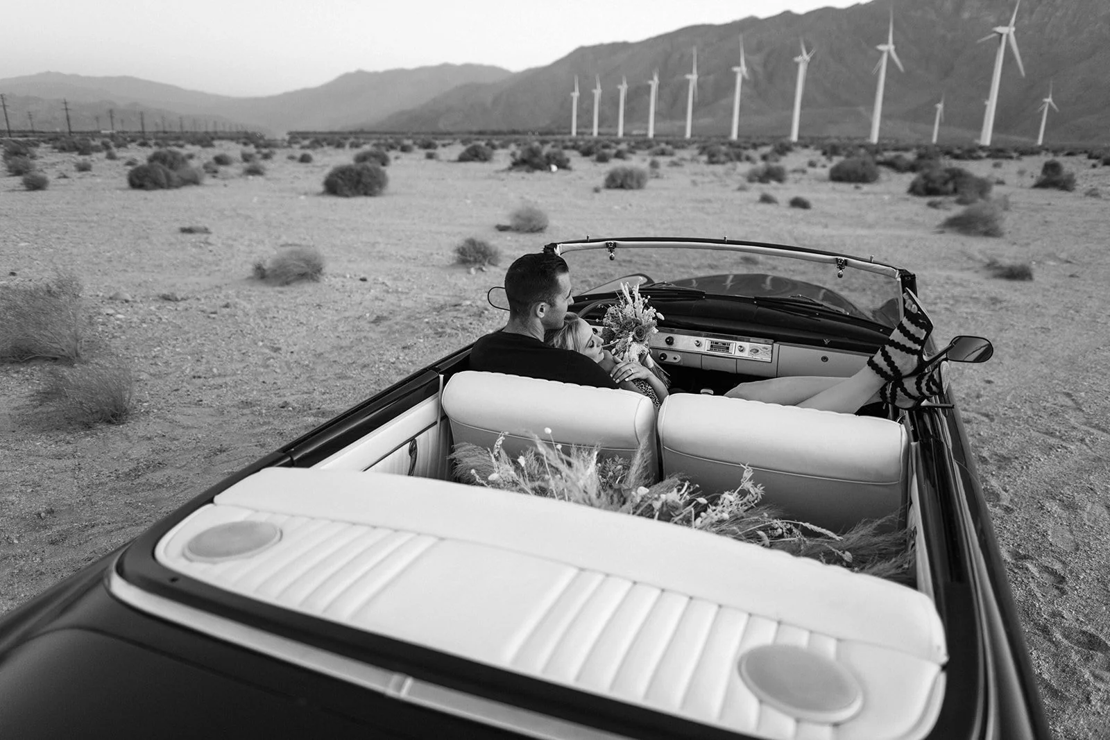 A black and white photo of a couple in a vintage convertible car in a desert landscape, with wind turbines and mountains in the background. The woman is lying with her legs over the dashboard, holding a bouquet, while the man sits beside her.