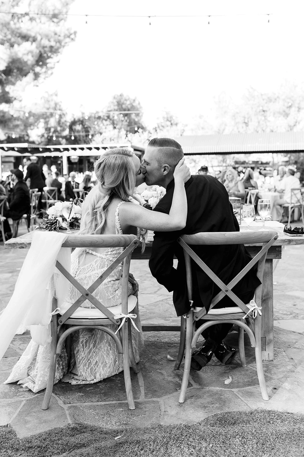 A black and white photo of a bride and groom sharing a kiss at their wedding reception, sitting at a wooden table outdoors with guests and string lights in the background.