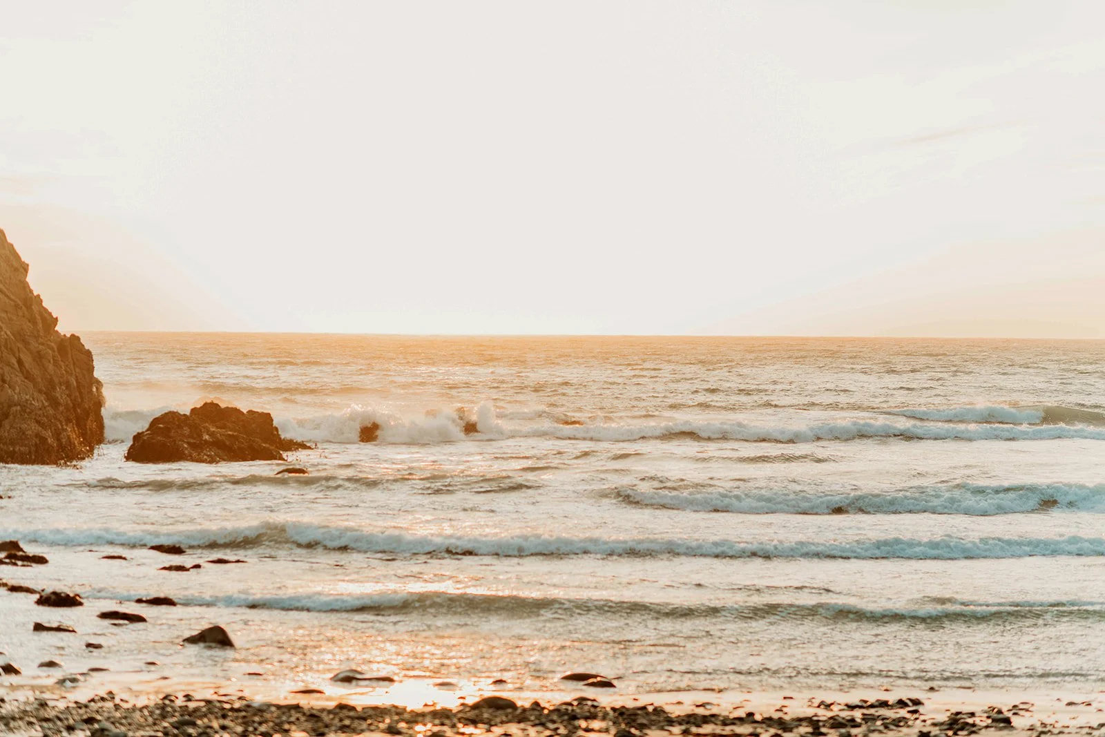 A serene beach scene during sunset with ocean waves crashing against rocks, a sandy shore with pebbles, and a partly cloudy sky.