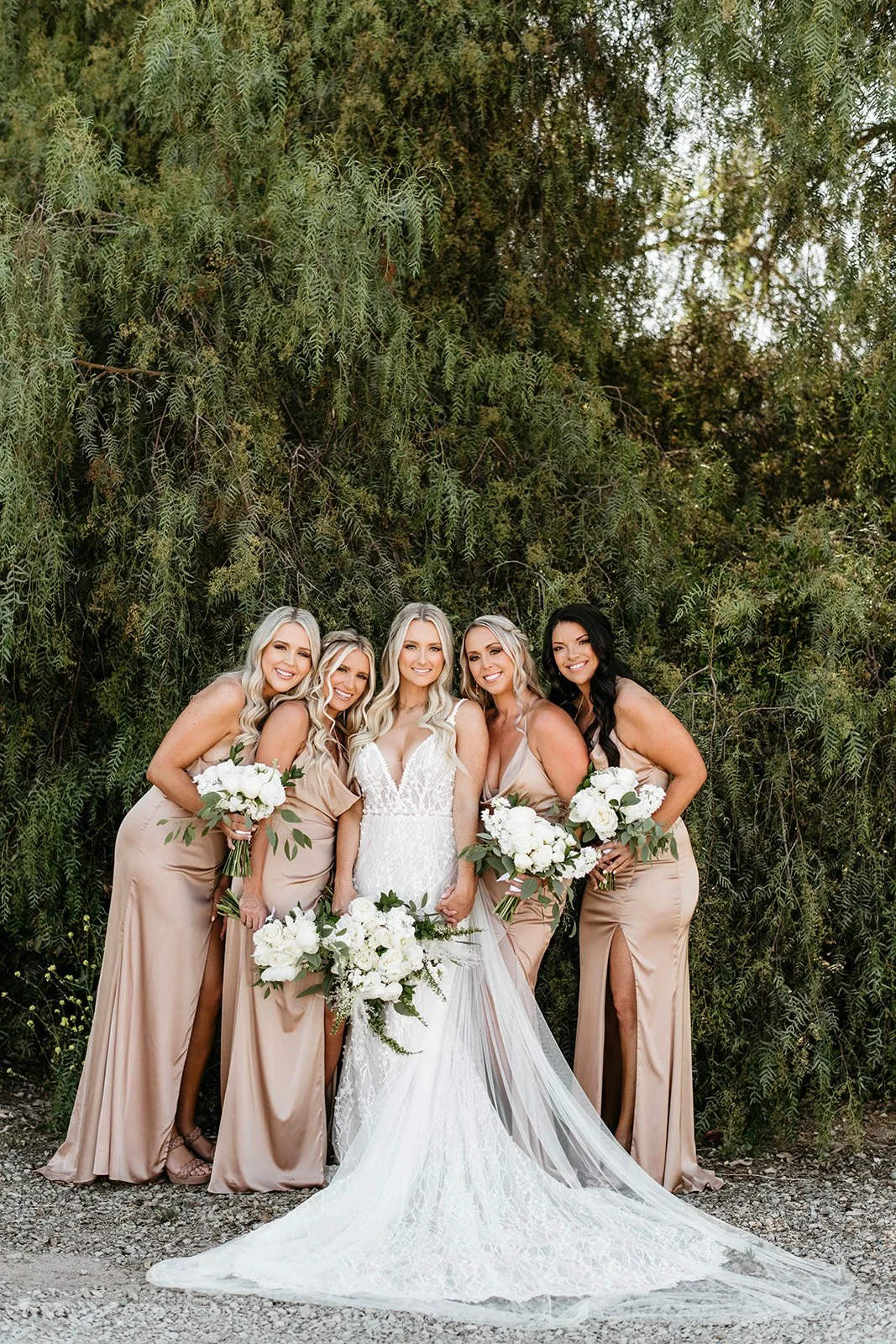 A bride in a white lace wedding gown holding a bouquet standing with four bridesmaids in beige dresses holding bouquets, in front of a lush green background.