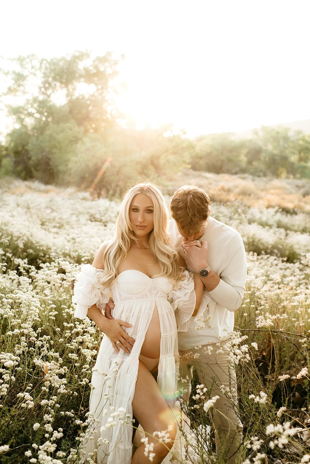 A pregnant woman and a man in a field of white flowers at sunset. The woman is wearing a white dress and the man is in white clothing, and he is kissing her hand.