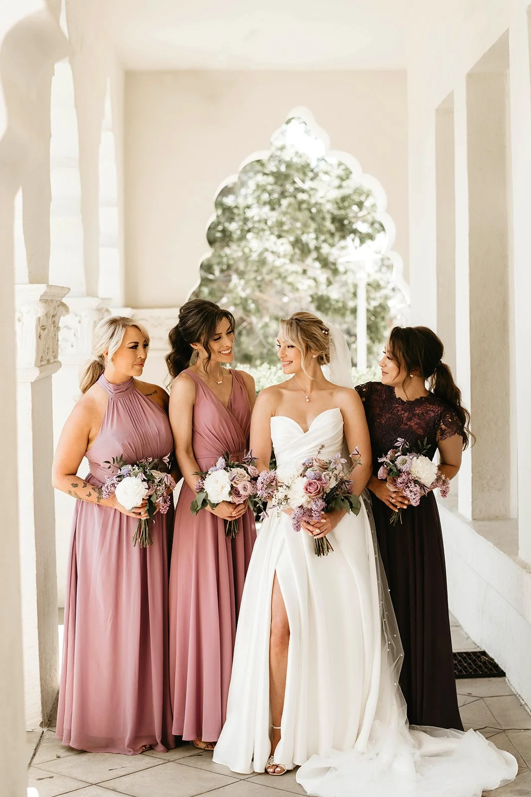 Bride and three bridesmaids standing outdoors under a stone archway, all holding bouquets of flowers, with a green tree in the background.