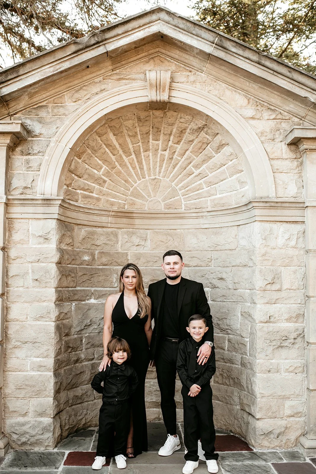 A family of four dressed in black, standing in front of a stone building with an arched window. The family includes a woman, a man, and two young boys.