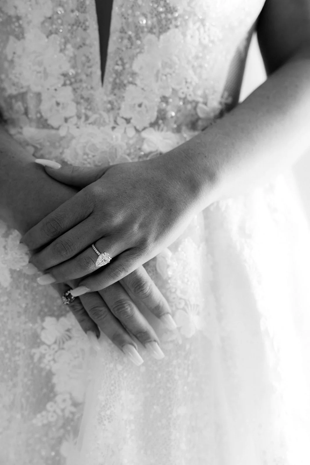 Close-up of a woman’s hands wearing a wedding ring, resting on a lace dress fabric, in black and white.