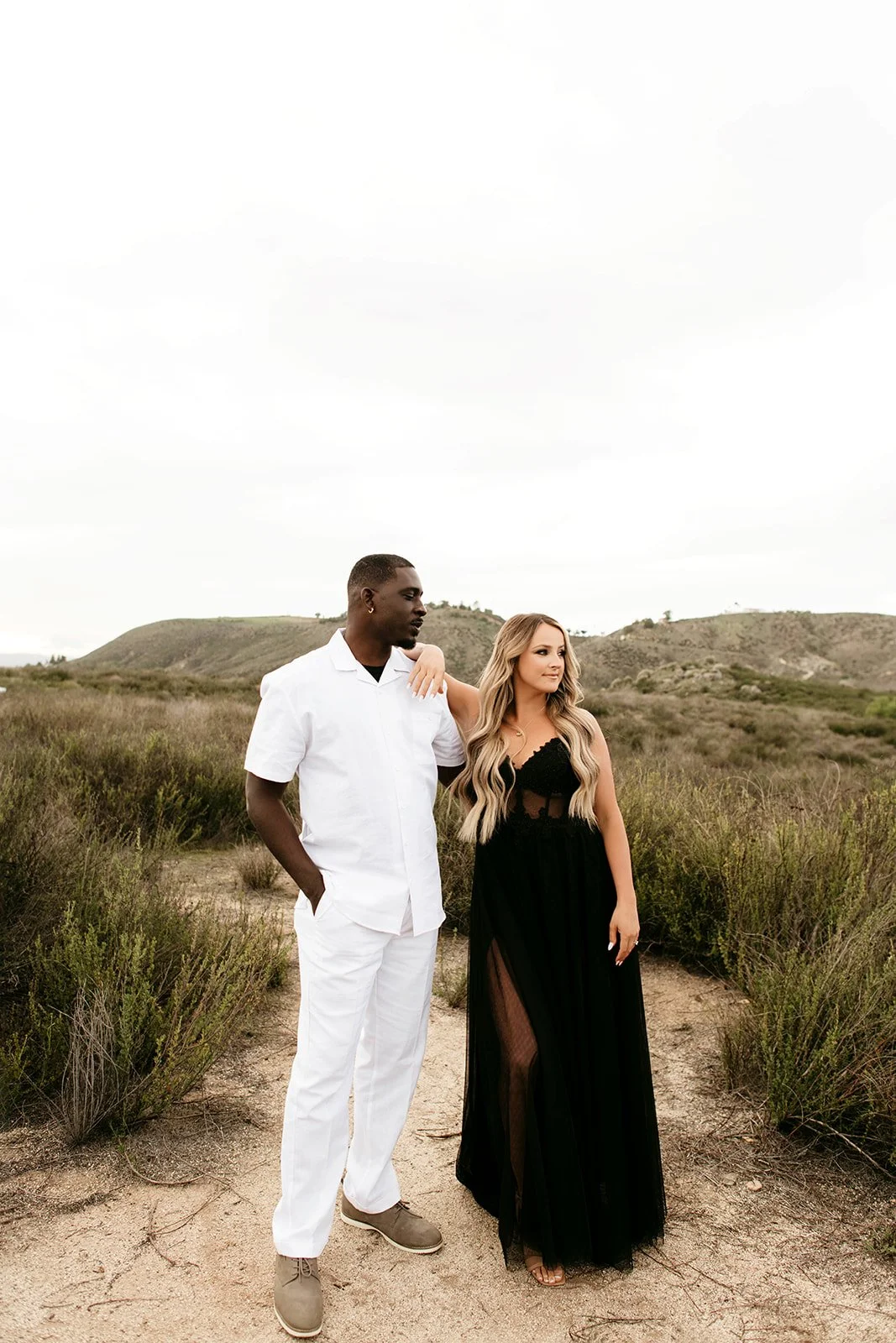 A man and a woman stand close together in a natural outdoor setting with rolling hills and grass, the woman arm draped over the man's shoulder, dressed stylishly, the man in a white shirt and pants, both gazing into the distance.