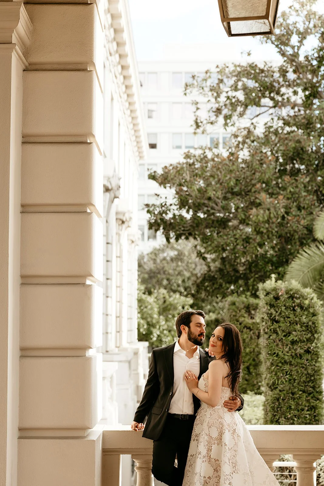 A couple in wedding attire standing on a balcony with a cityscape background and greenery.