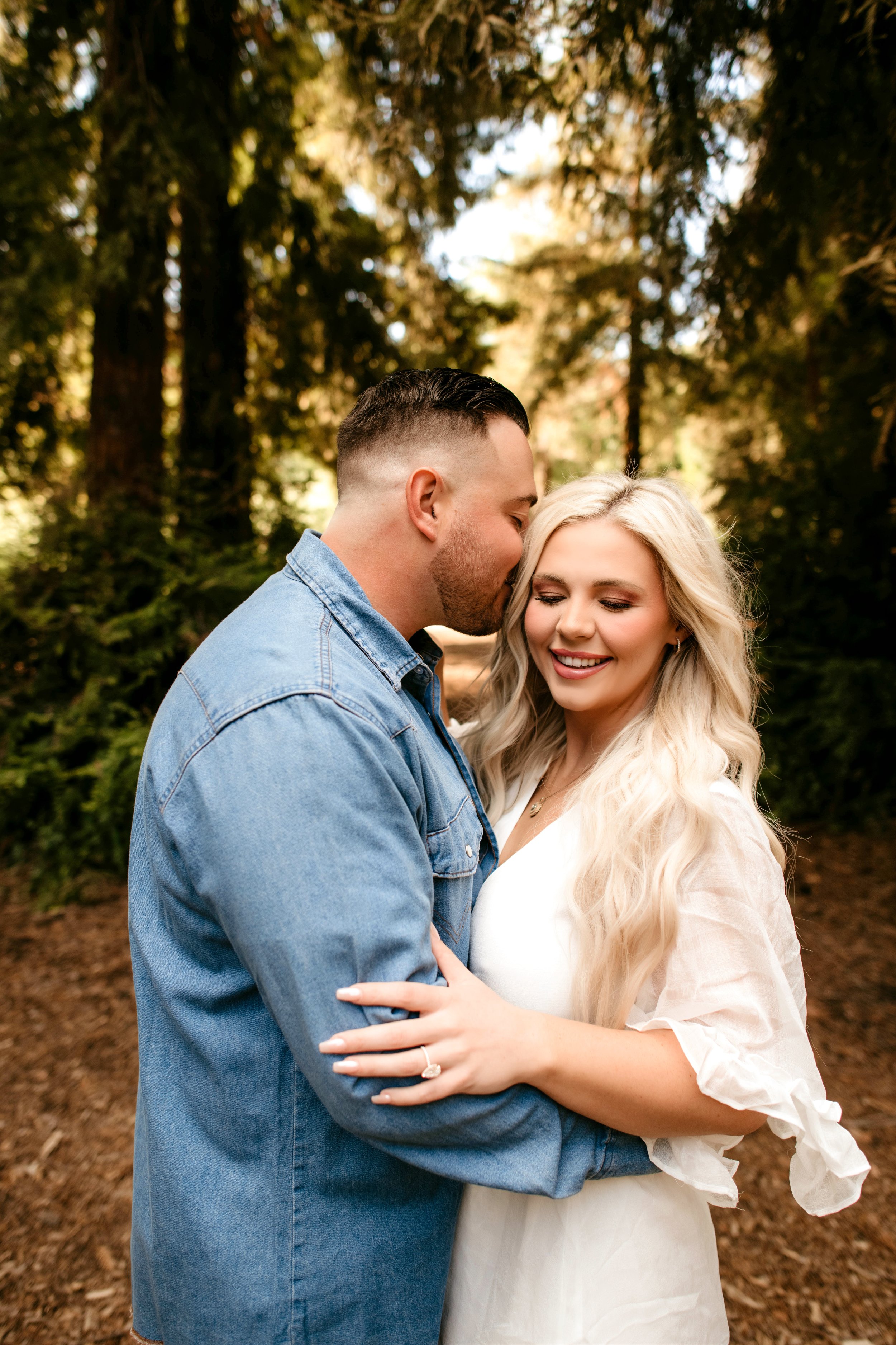 A man and woman embracing outdoors in a forest setting, looking happy and affectionate.