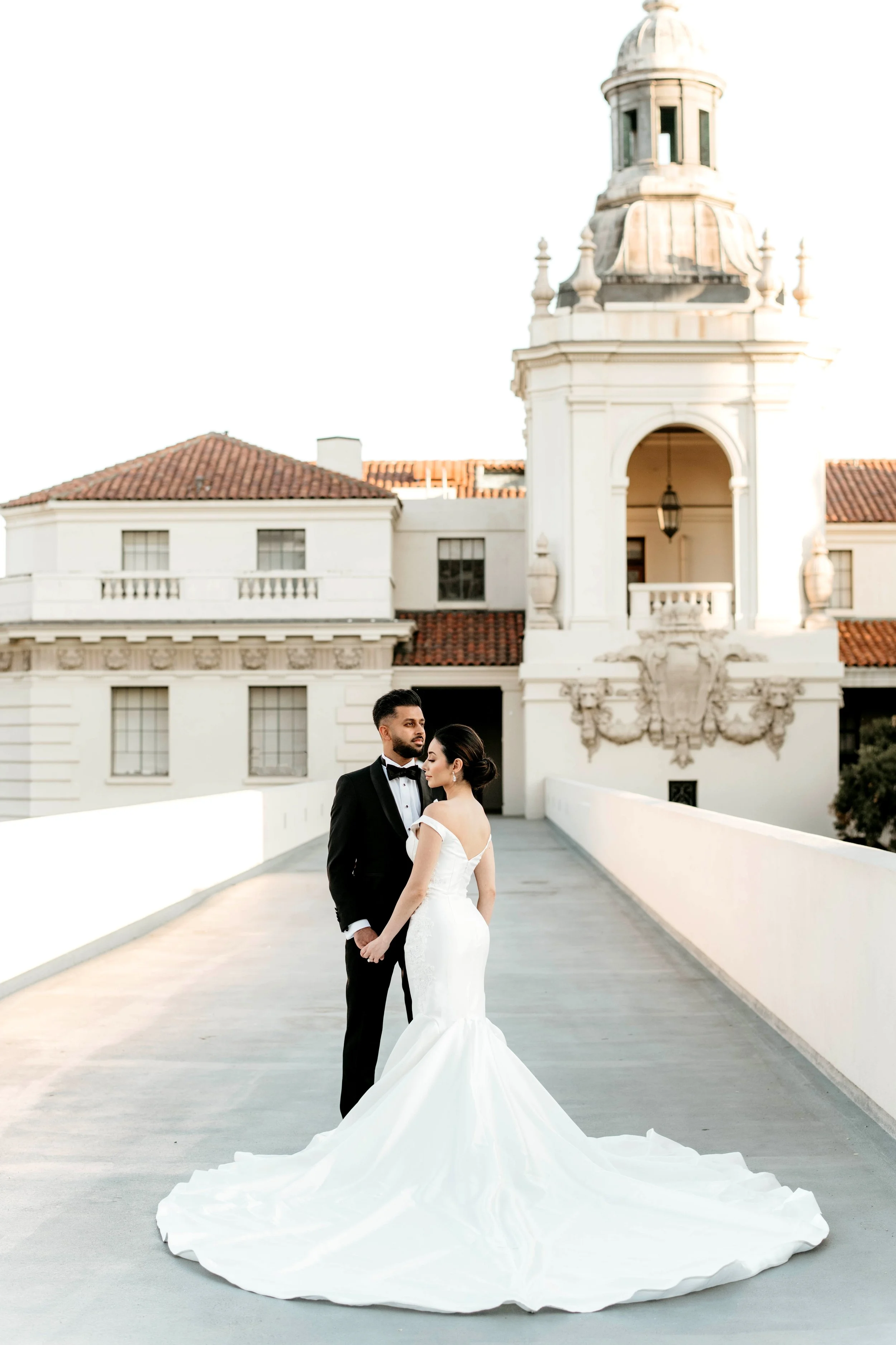 A bride and groom stand together on a white bridge in front of a historic building with a red-tiled roof and ornate architectural details. The bride wears a strapless white wedding gown with a long, flowing train, and the groom wears a black tuxedo w