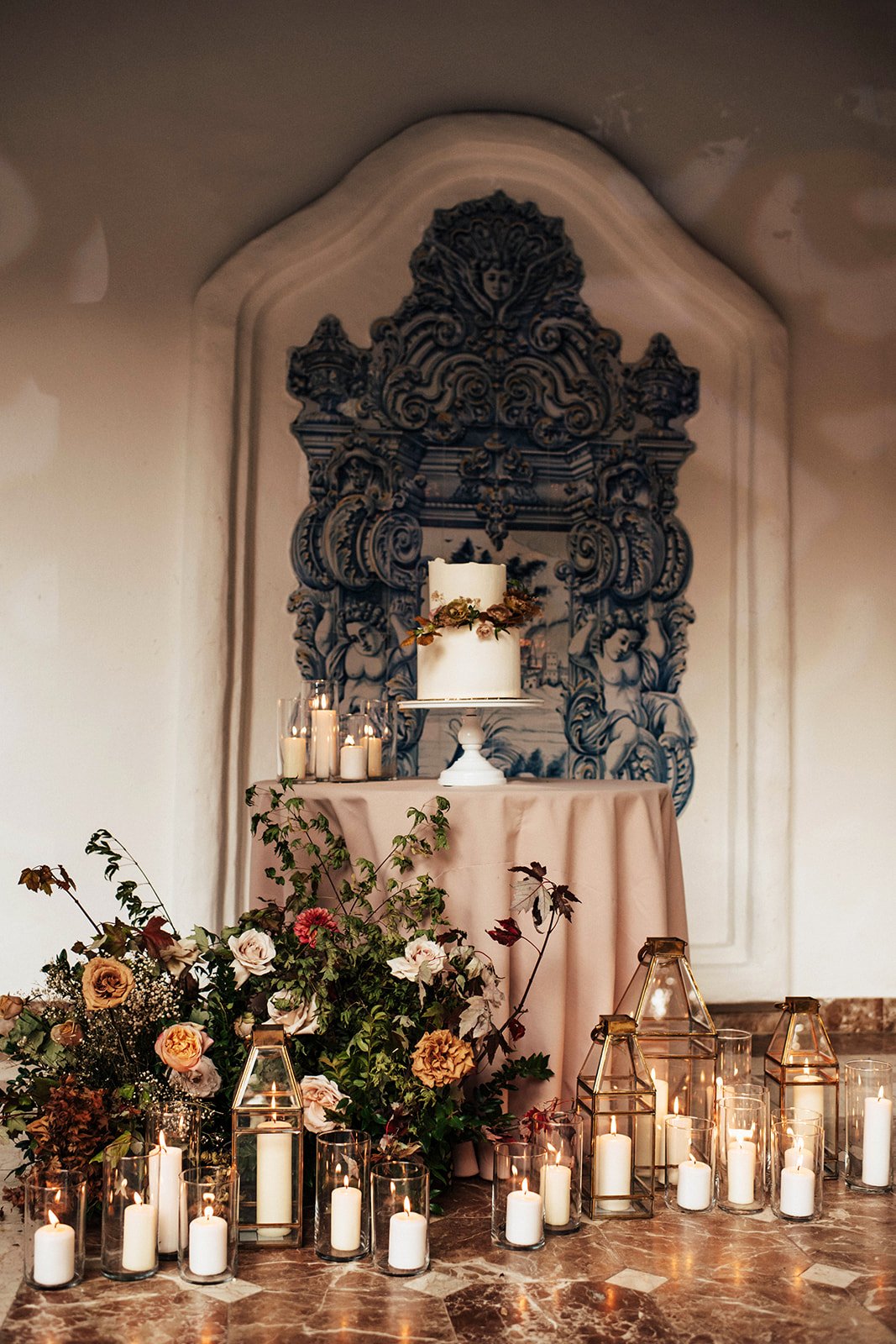 Elegant wedding display with a white cake adorned with flowers on a white stand, surrounded by candles in glass holders and lanterns, and floral arrangements in front, in front of a mural depicting cherubs and ornate designs.