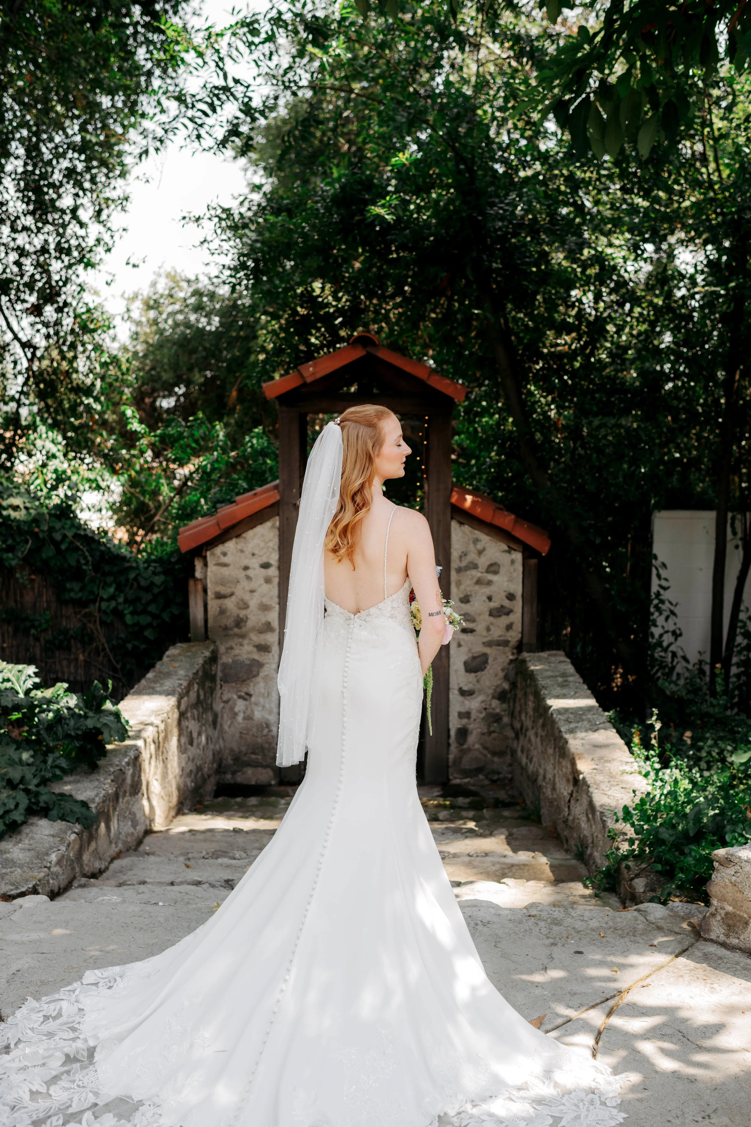 A bride in a white wedding dress with lace details and a long veil stands in an outdoor garden setting with greenery and stone structures, facing sideways.