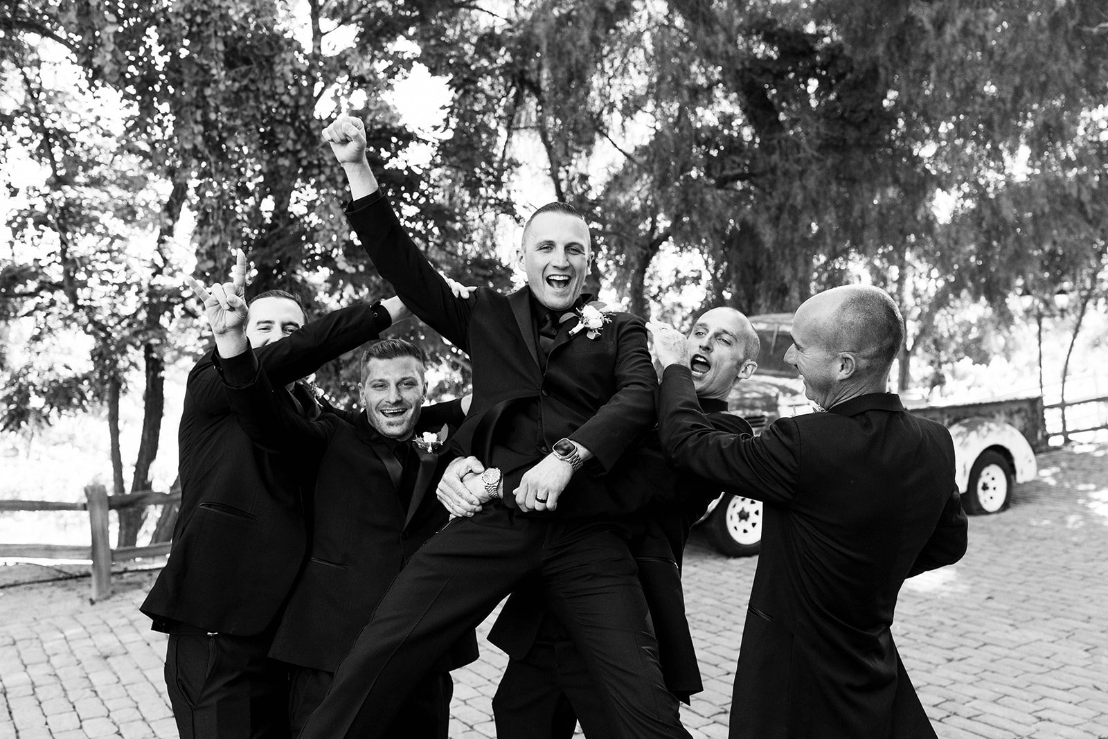 A group of five men dressed in black suits, celebrating outdoors. One man is being lifted by the others, all smiling and showing excitement. The background has trees, a wooden fence, and a parked white car.