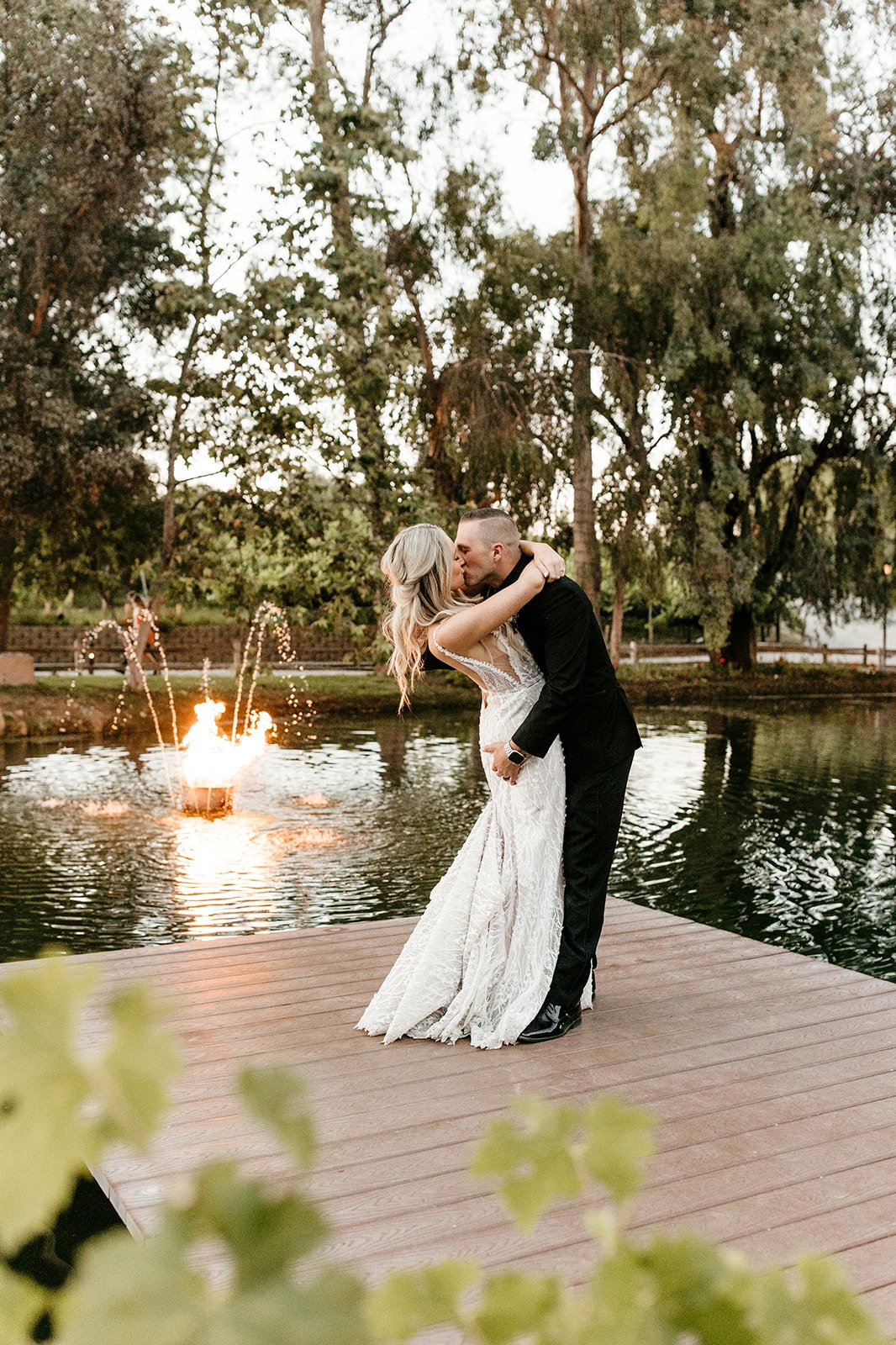 A couple in wedding attire sharing a kiss on a wooden dock near a pond, with a small fire and sparklers in the background, surrounded by trees.