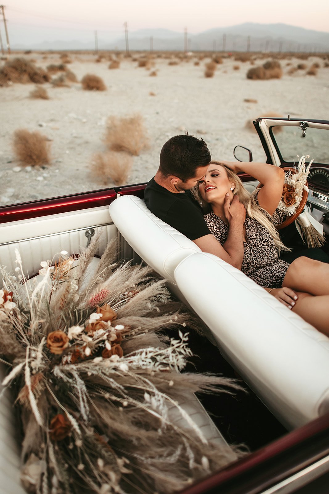 Couple sitting in a vintage car with desert landscape in the background, decorated with flowers and dried plants.