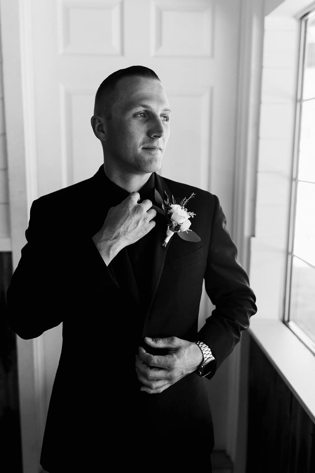 Black and white photo of a man in a suit adjusting his boutonniere, looking out the window.